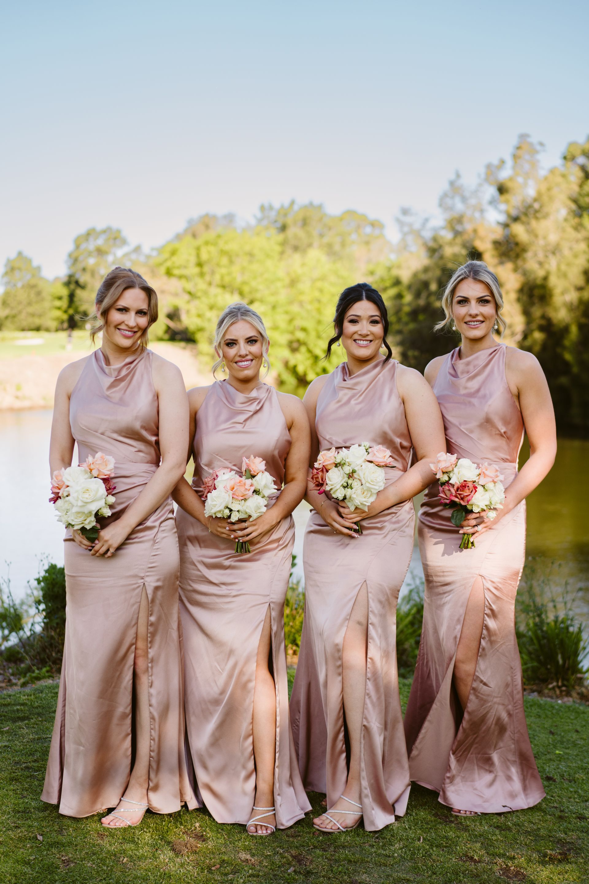 Four bridesmaids in blush dresses holding bouquets outdoors on a sunny lawn
