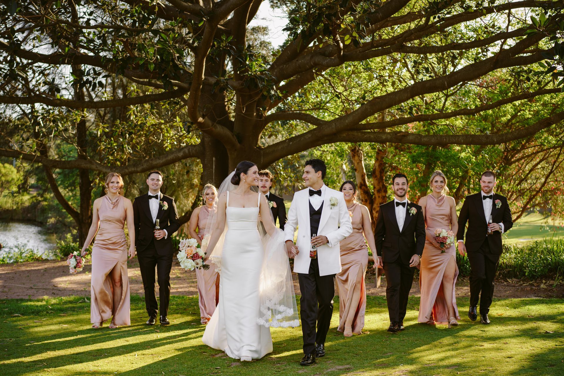 Wedding party walking on a grassy lawn under a large tree, with bride in white and attendants in blush and black suits