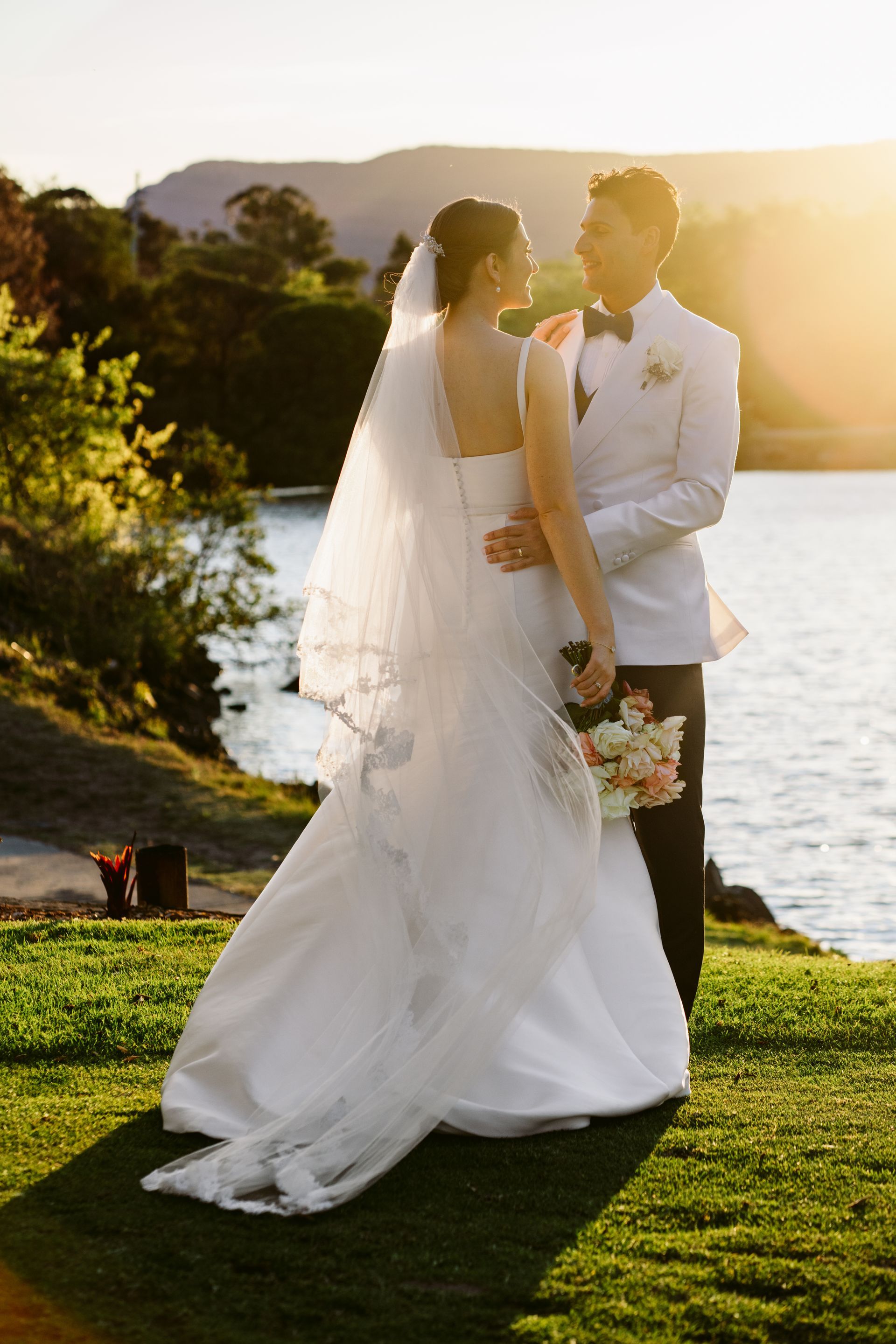 Bride and groom embracing by a lake at sunset, with mountains and a bouquet visible