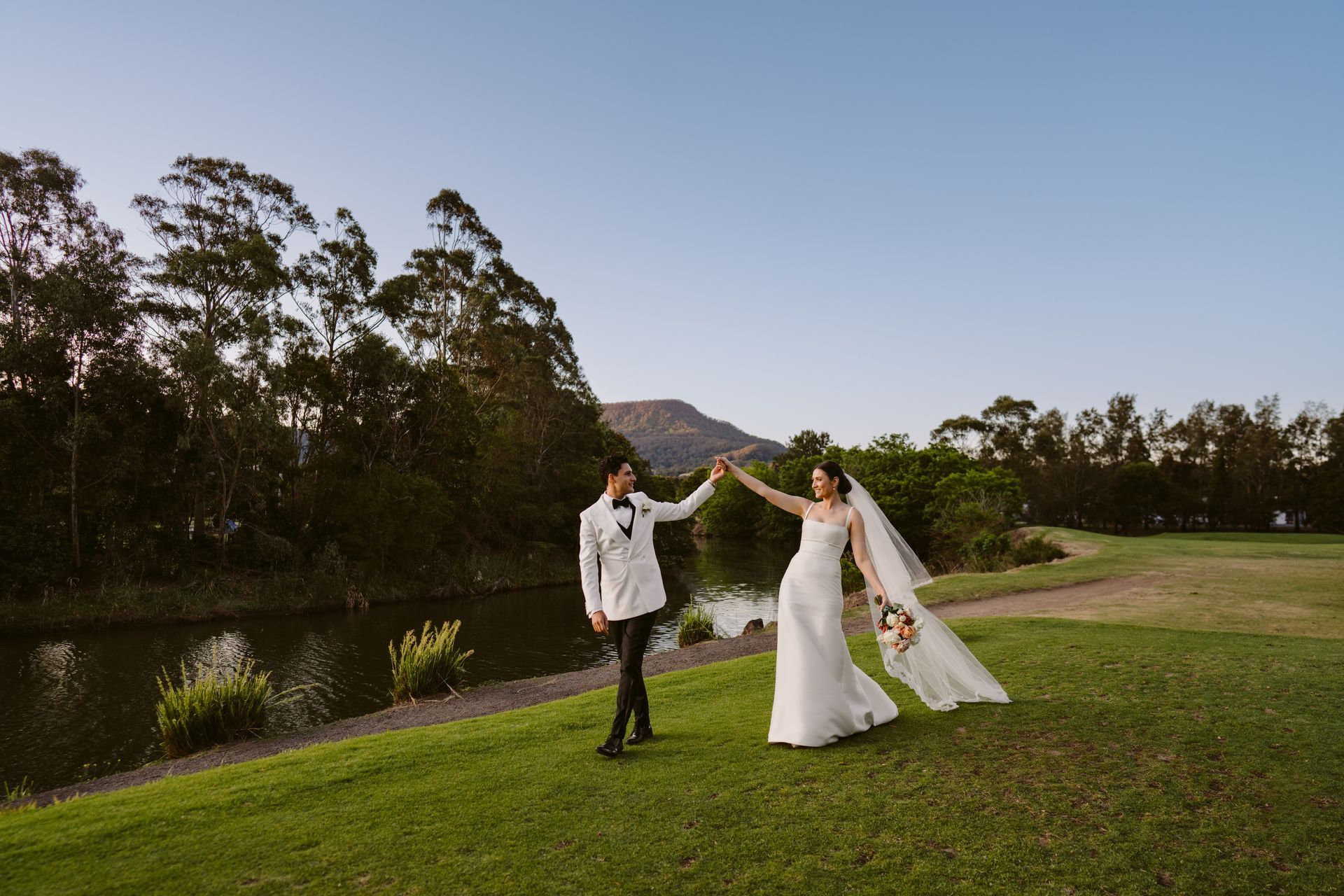 Bride and groom in white dancing on a grassy lawn by a river at sunset