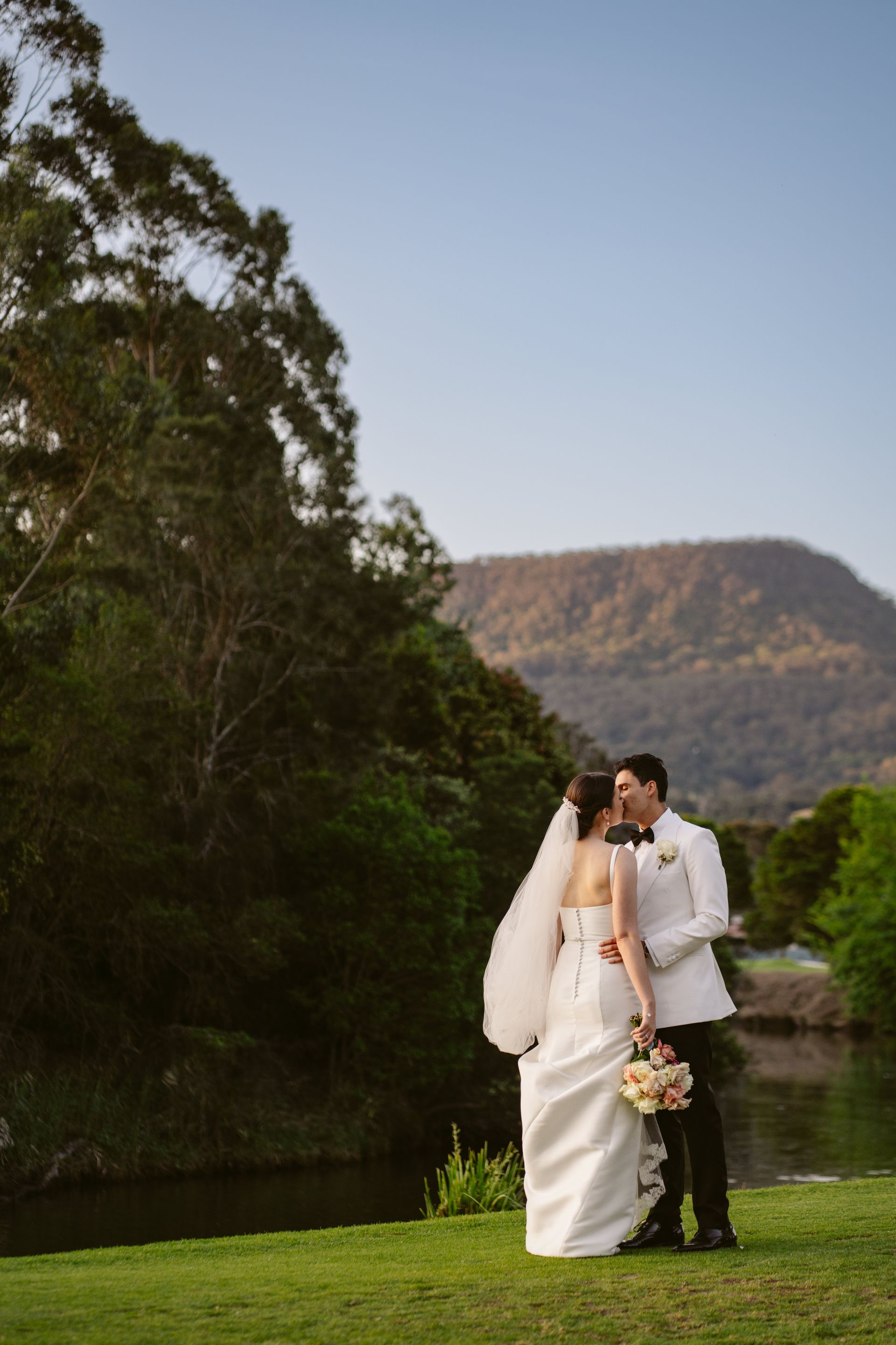 Bride and groom in white wedding attire embracing outdoors by a lake and mountain backdrop