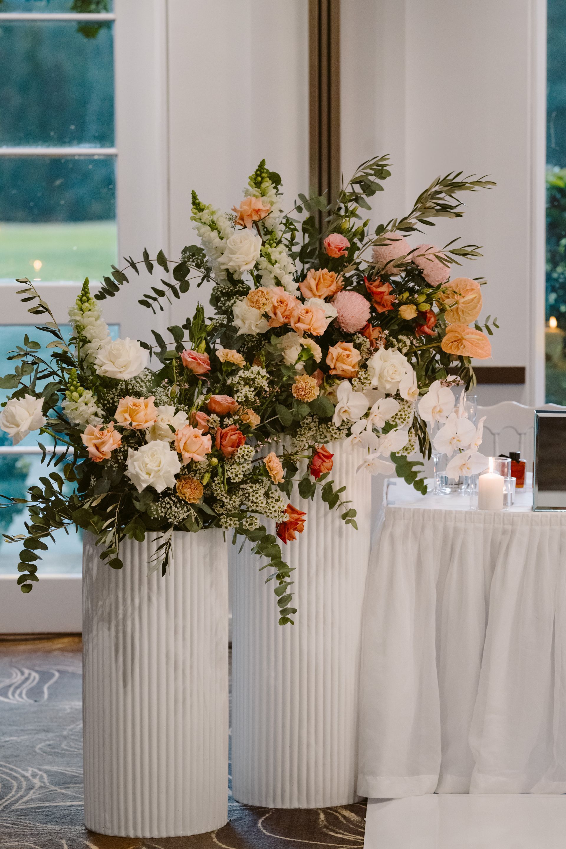 Elegant floral arrangement with peach and white flowers on tall ribbed pedestals beside a draped table