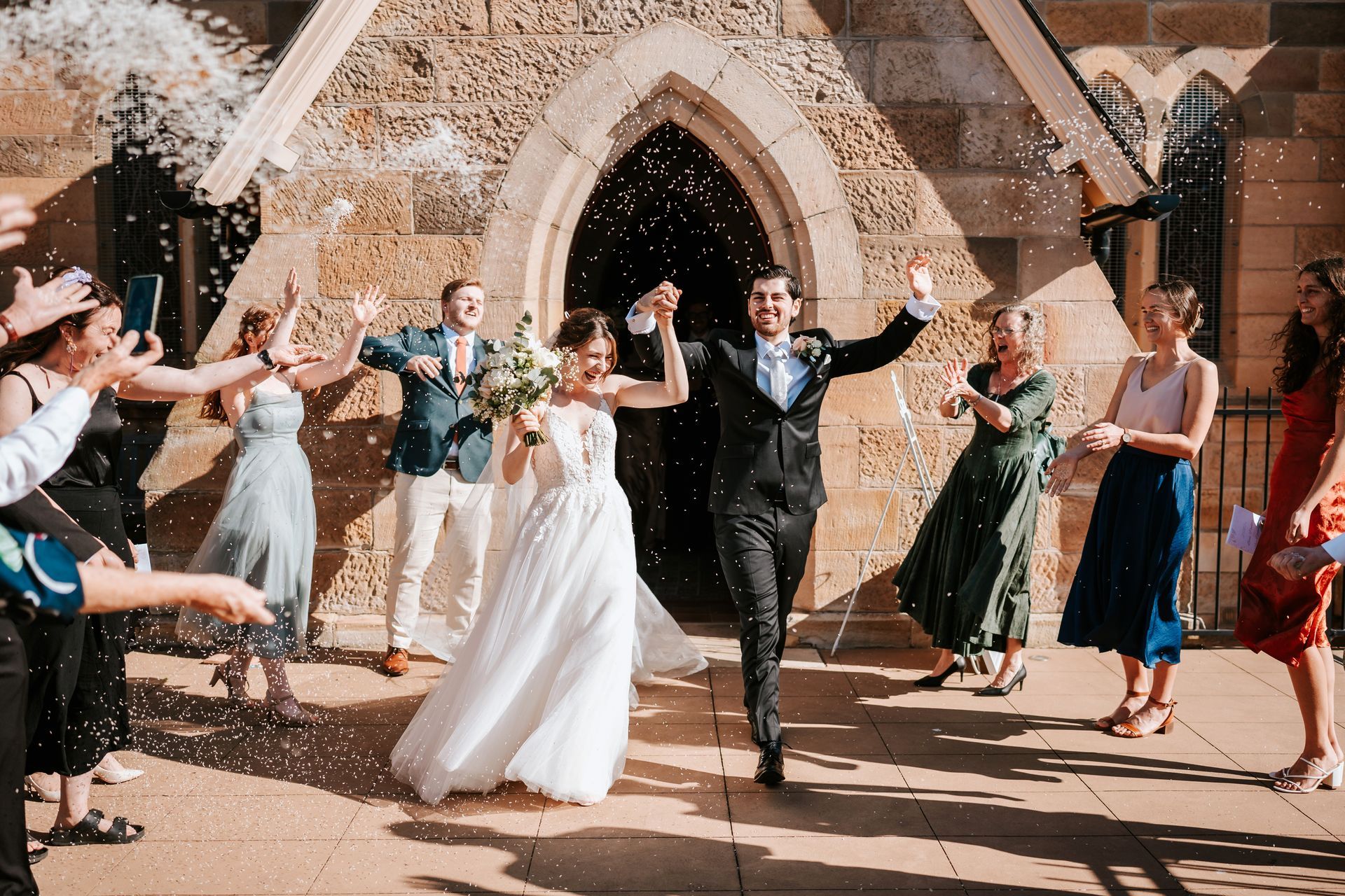 Wedding couple walking through an arch of confetti outside a stone church, guests cheering around them