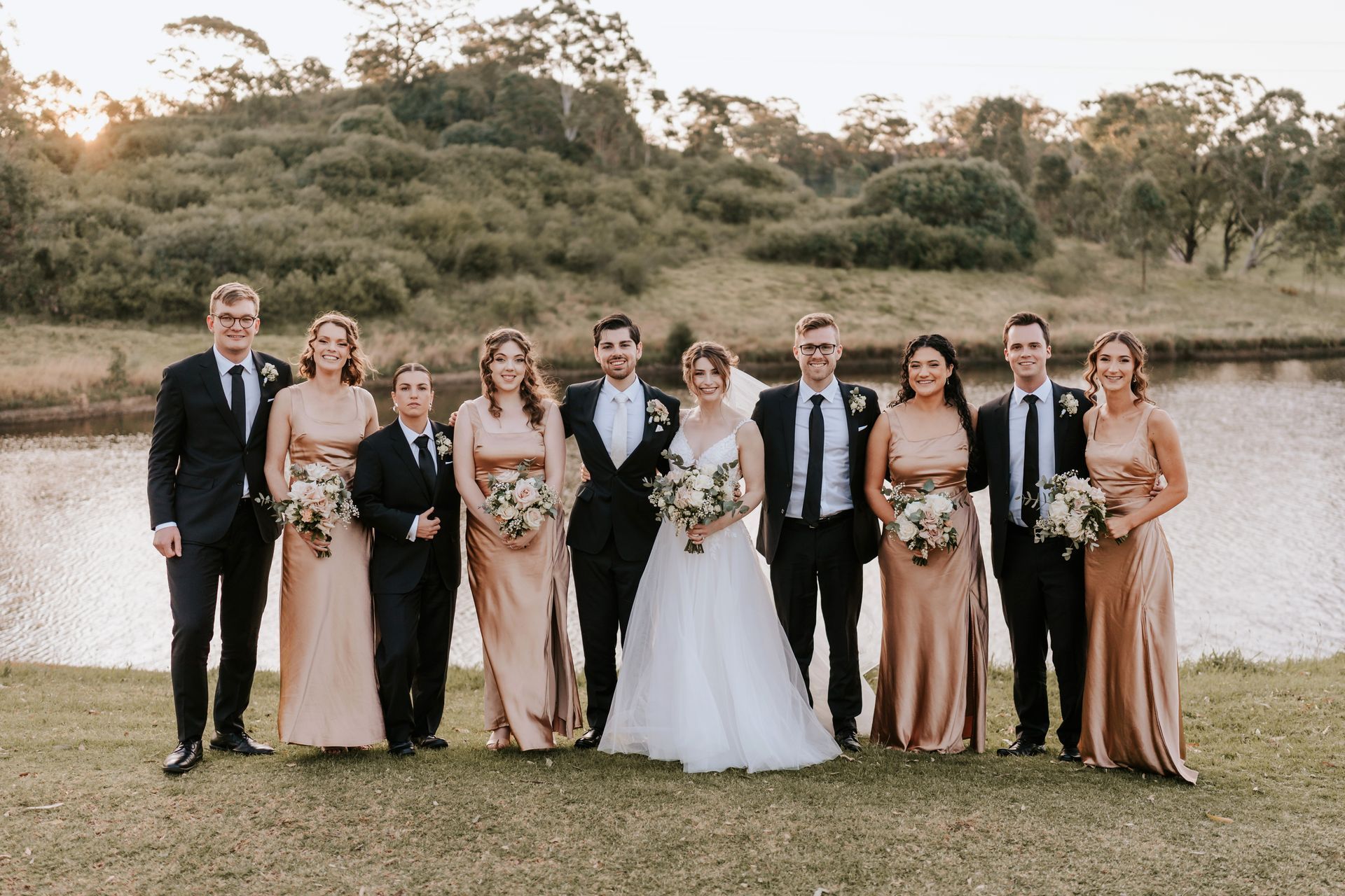 Wedding party standing outdoors by a river, with the bride in a white gown and guests in formal attire
