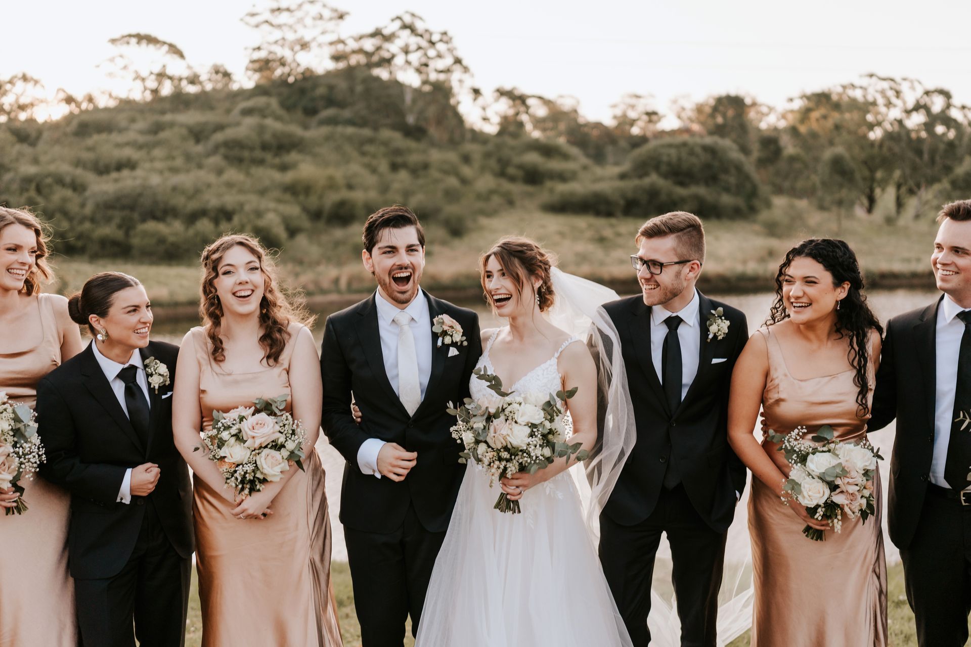 Wedding party standing outdoors in formal attire, holding bouquets and smiling at the camera.