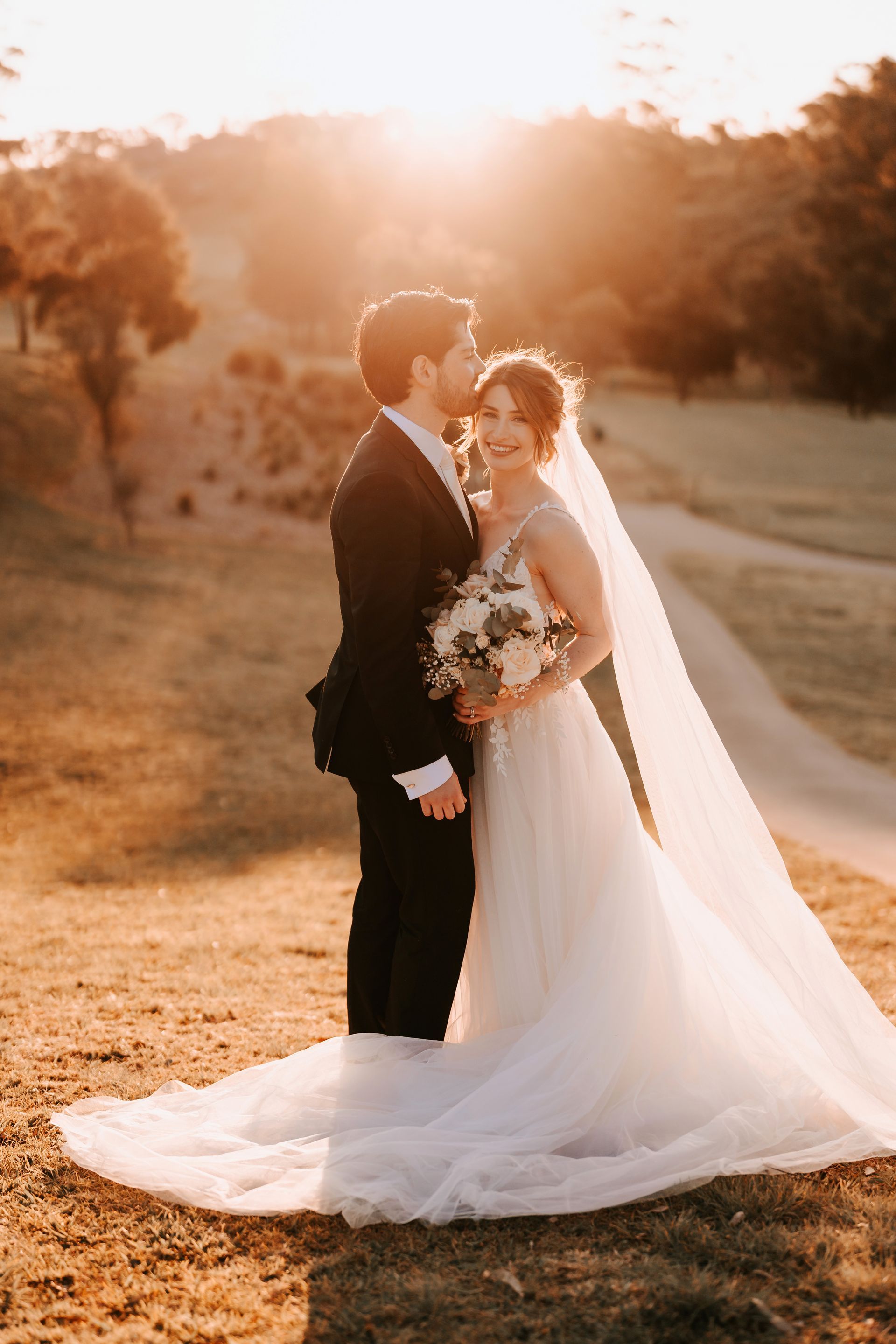 Bride and groom embracing outdoors at sunset, with the bride’s long white veil flowing behind her