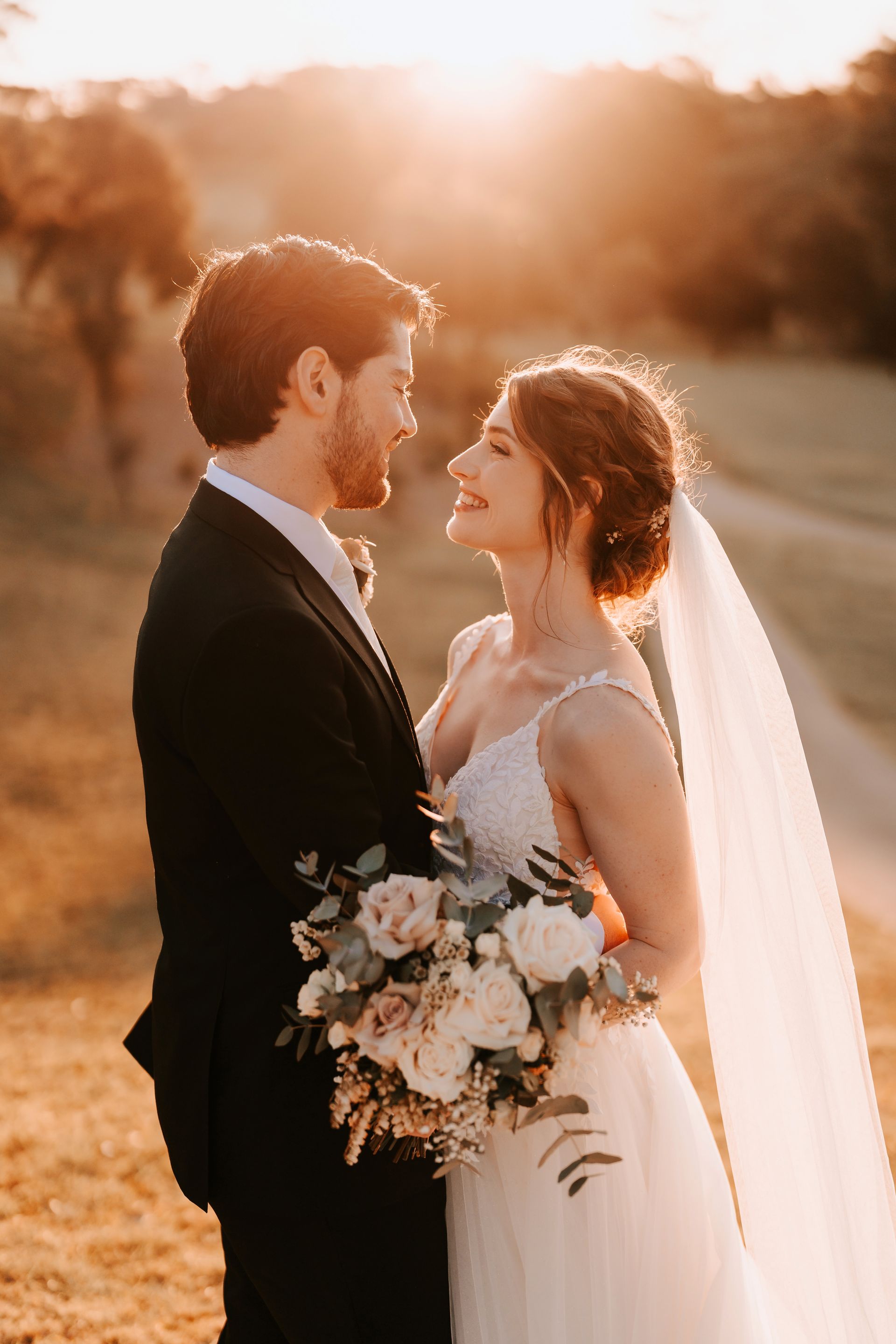 Bride and groom face each other outdoors at sunset, with warm backlight and a bouquet in the bride’s hands