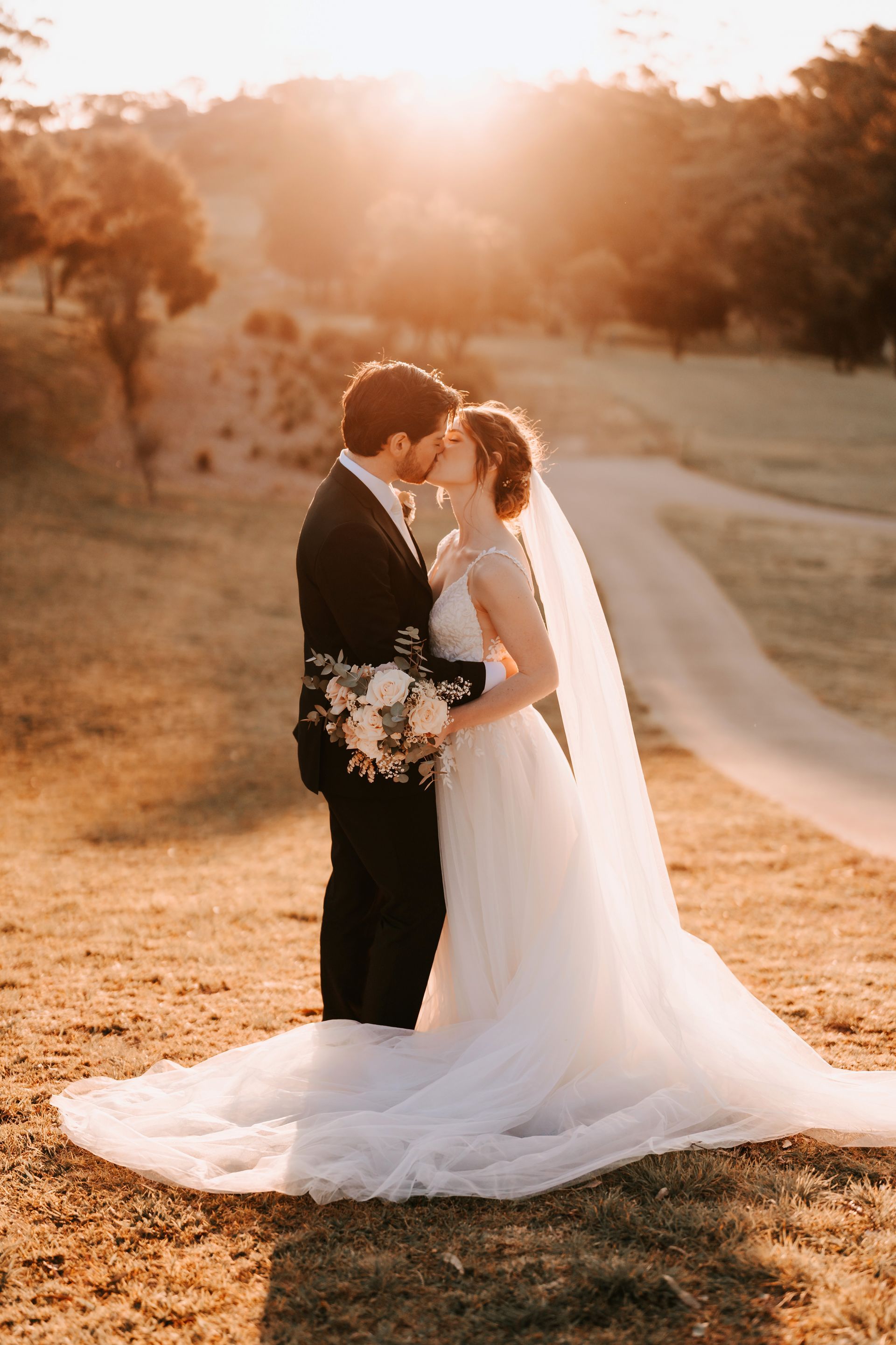 Bride and groom kiss at sunset in a golden field, bride in a long white gown with veil