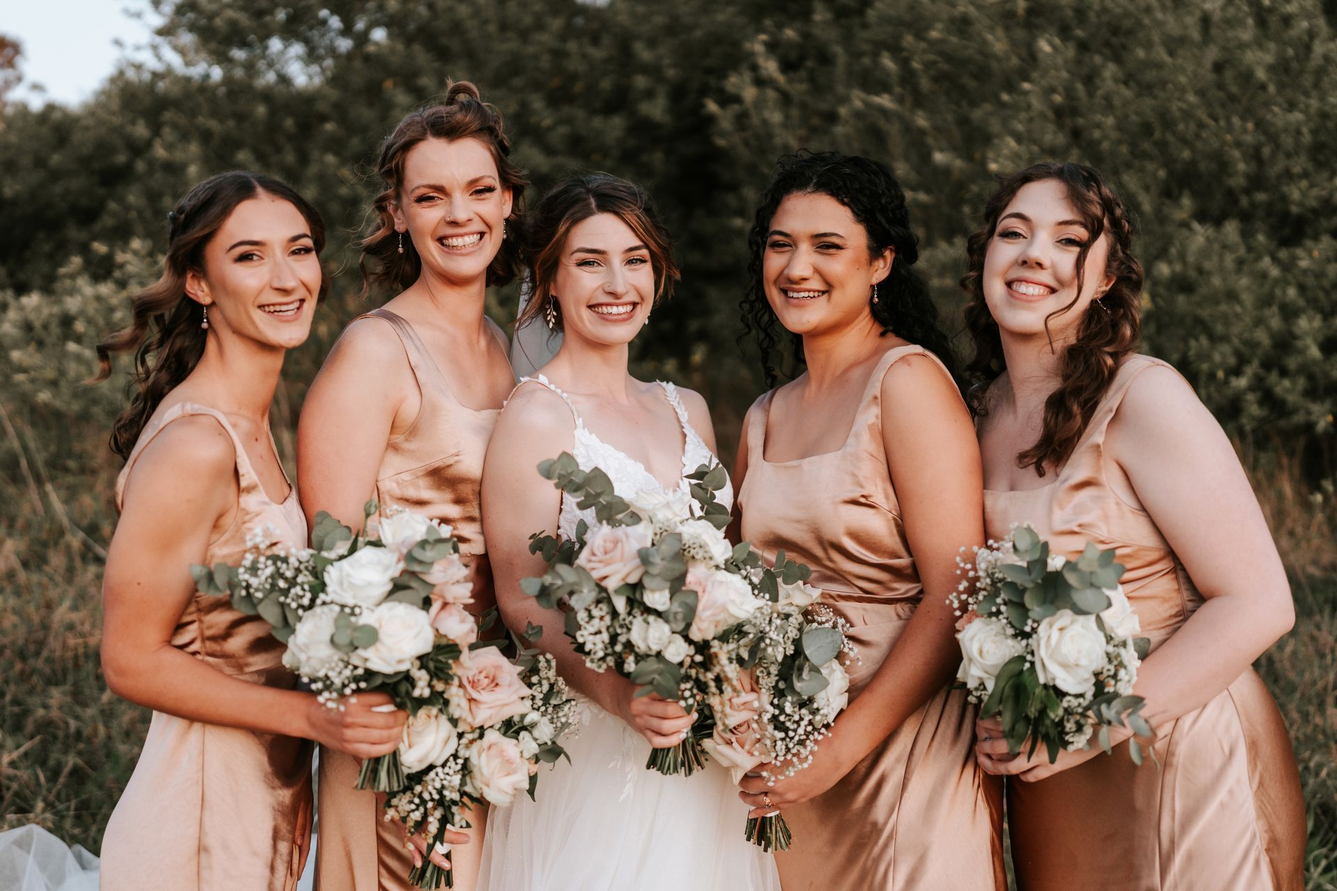 Five bridesmaids in blush dresses holding white bouquets and smiling outdoors
