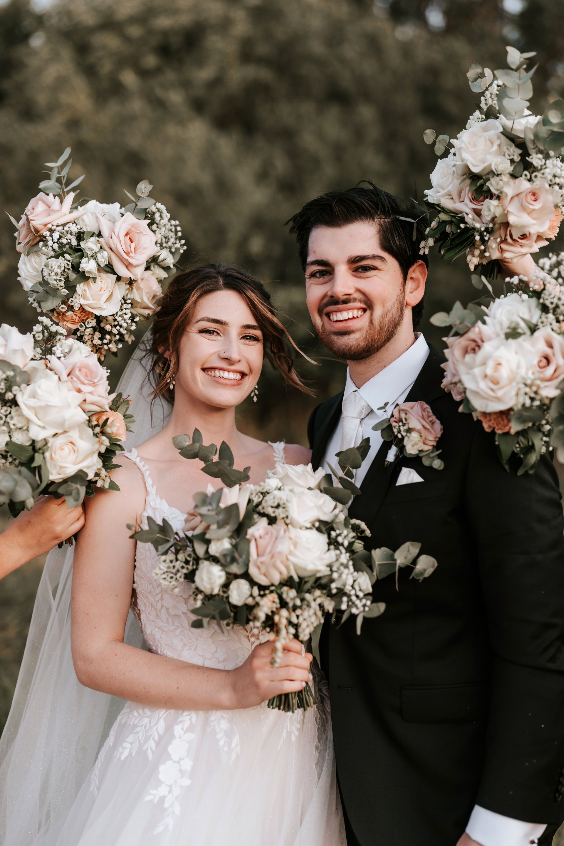 Wedding couple holding bouquets, smiling outdoors with floral arrangements and greenery.