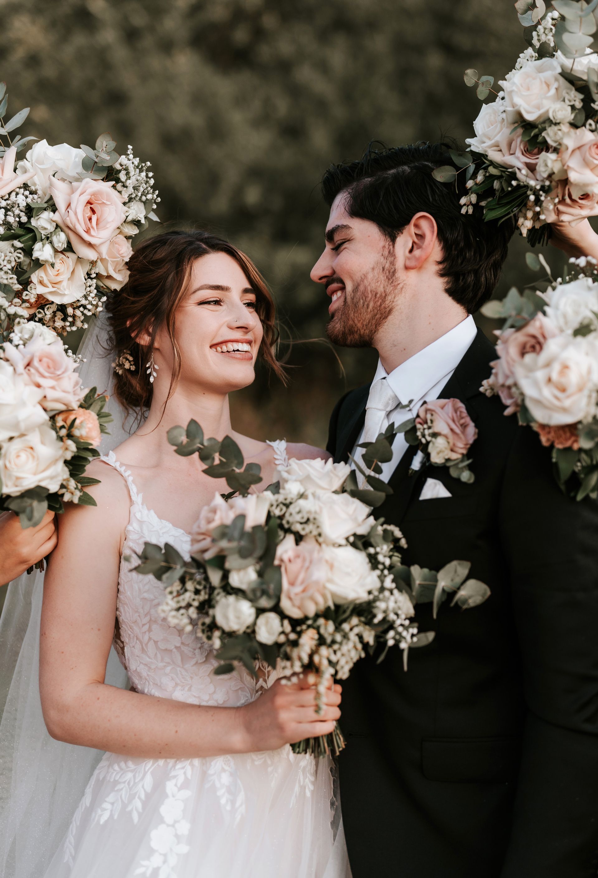 Wedding couple smiling under floral arch, holding bouquet in soft, romantic outdoor setting
