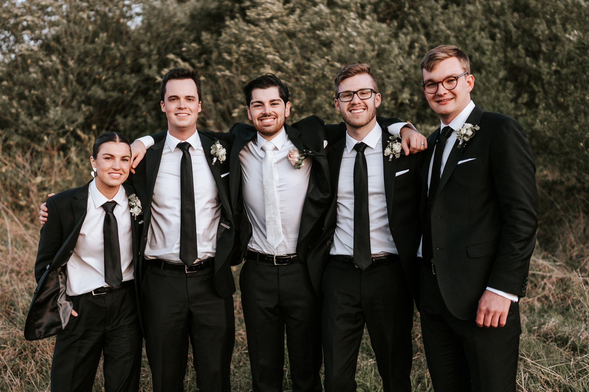 Five people in formal black suits standing together outdoors, smiling in front of trees.