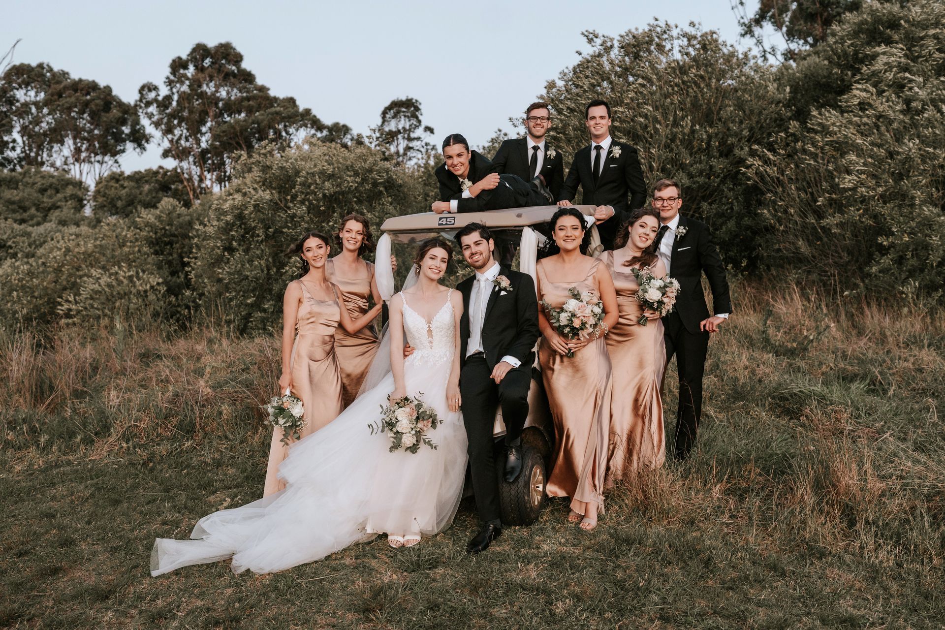 Wedding party posing outdoors in formal attire, with the bride in a white gown and bouquet.