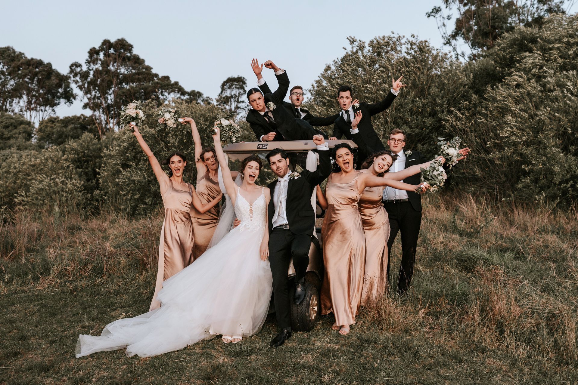 Wedding party posing outdoors, cheering with arms raised in a grassy field.