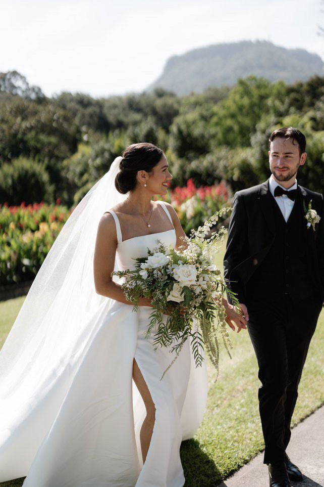Bride and groom in wedding attire walking outdoors with mountains and flowers in the background