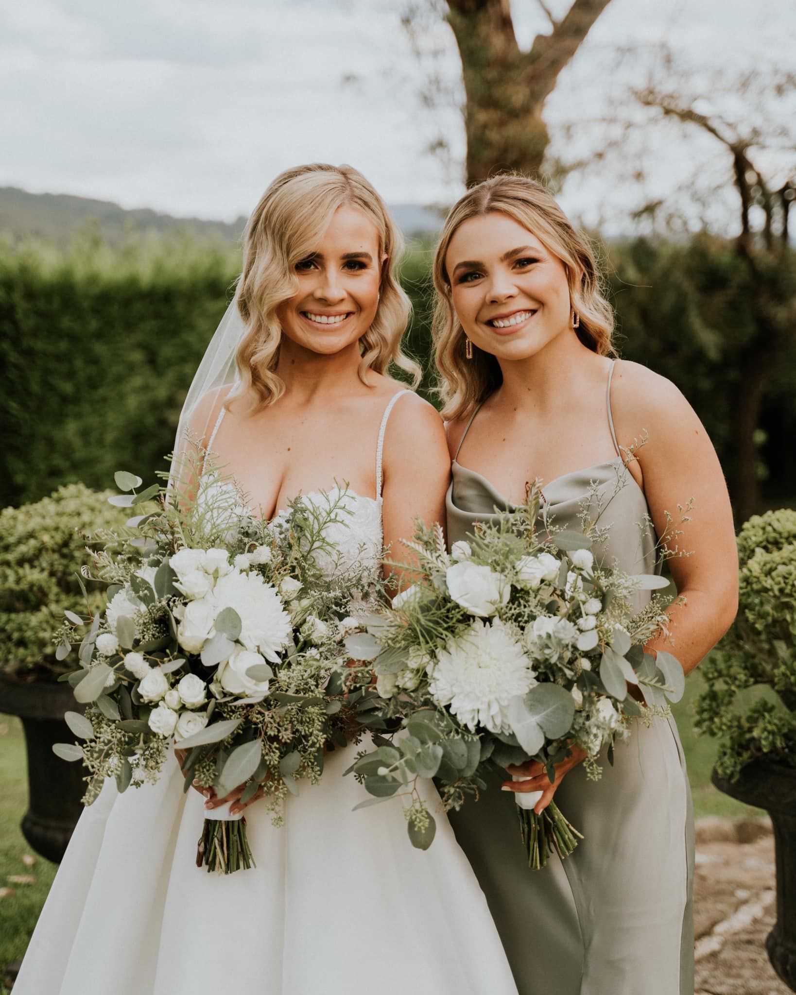 Two women in light dresses holding white bouquets outdoors, smiling beside each other.