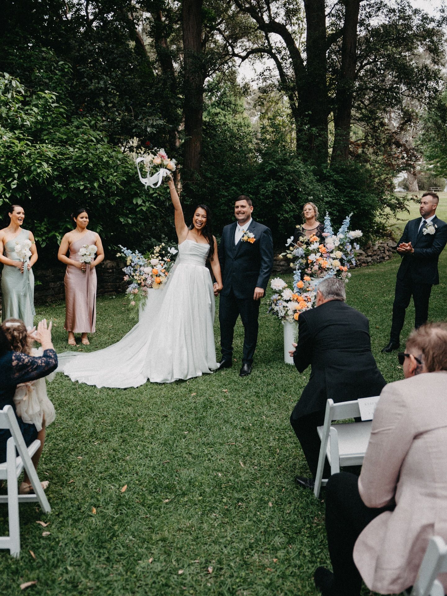 Wedding couple exchanging vows outdoors under trees, surrounded by attendants and seated guests.