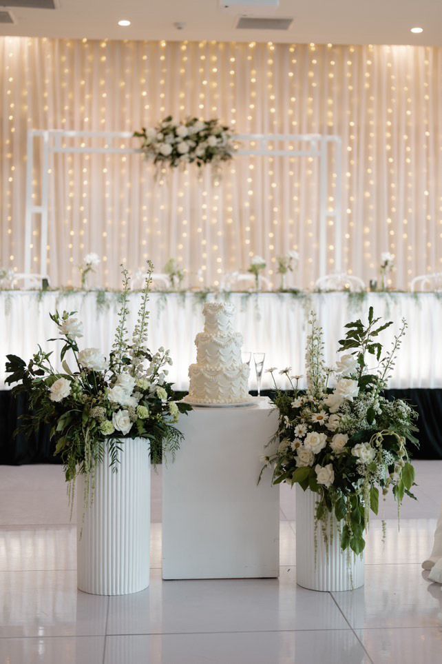 Elegant white floral wedding altar with draped lights and greenery arrangements on pedestals