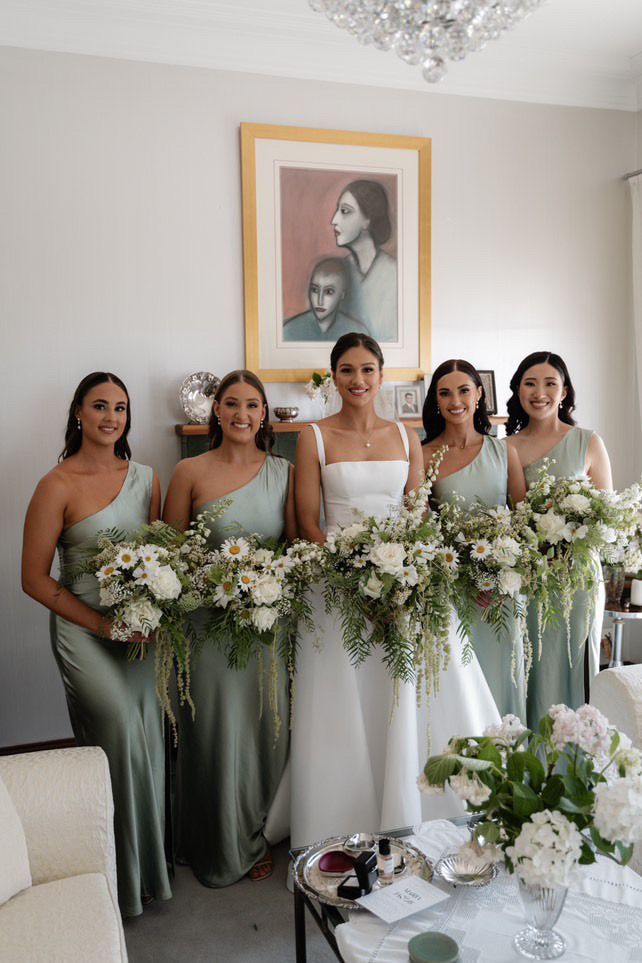 Five women in sage and white dresses holding white floral bouquets at an indoor wedding