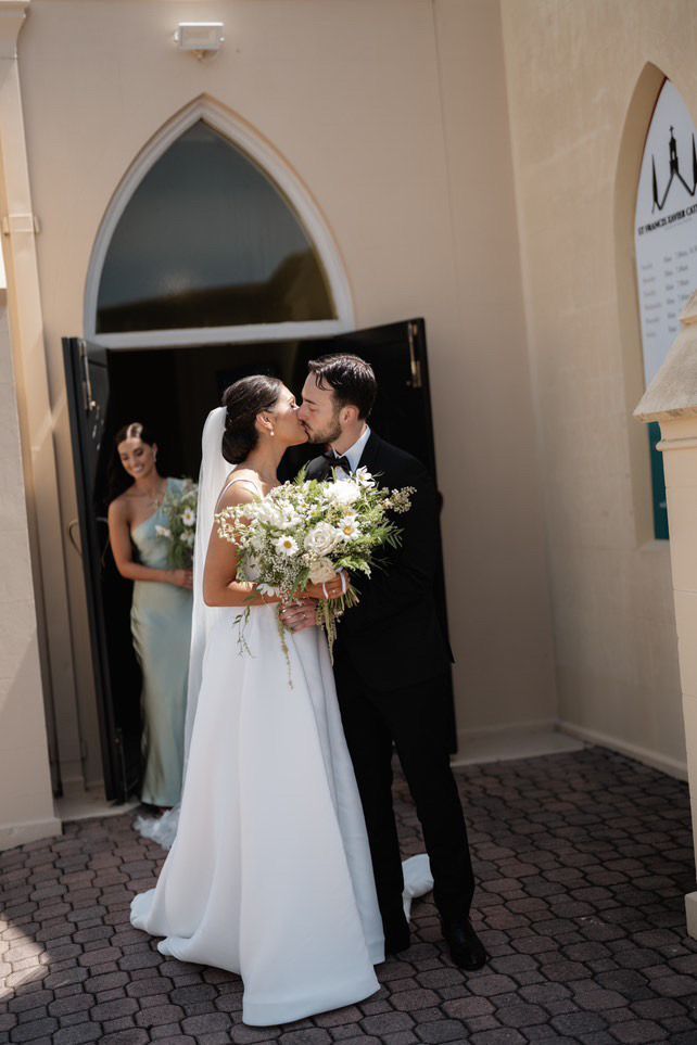 Bride and groom kiss outside a church doorway, bride holding a bouquet in a white dress