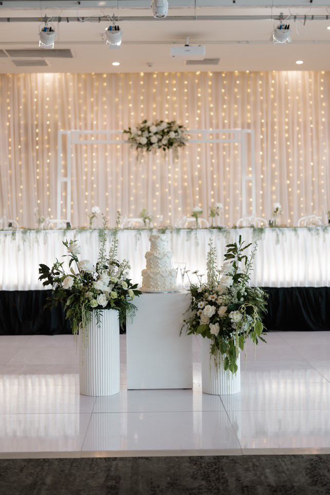Elegant wedding stage with white drapery, fairy lights, and tall floral arrangements on a glossy floor