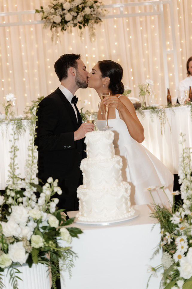 Wedding couple kissing behind a white tiered cake, with floral decor and warm string lights backdrop