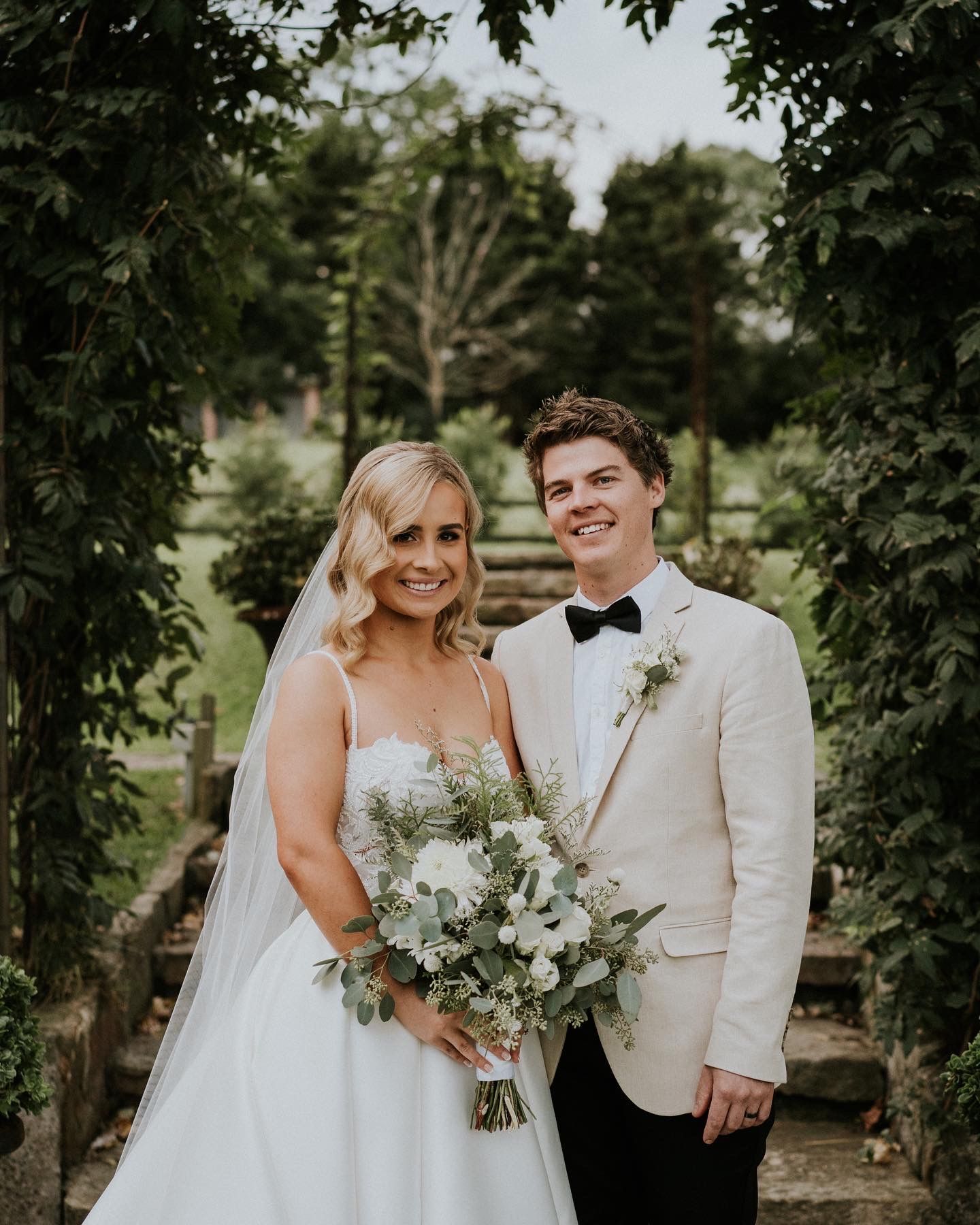 Bridal couple posing outdoors in wedding attire, with bouquet and trees in the background