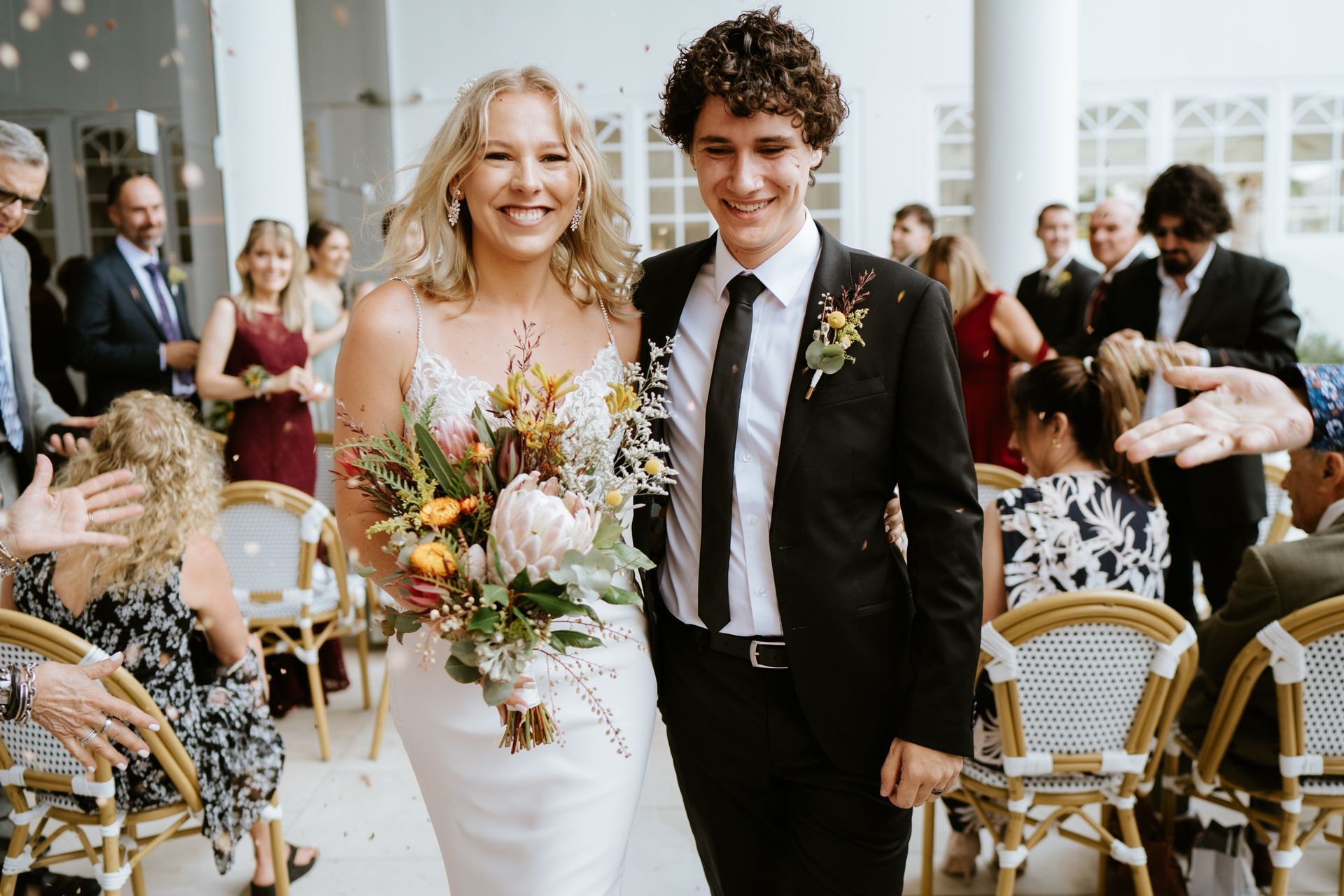 Wedding couple walking down the aisle, smiling, with guests seated in a bright ceremony hall.