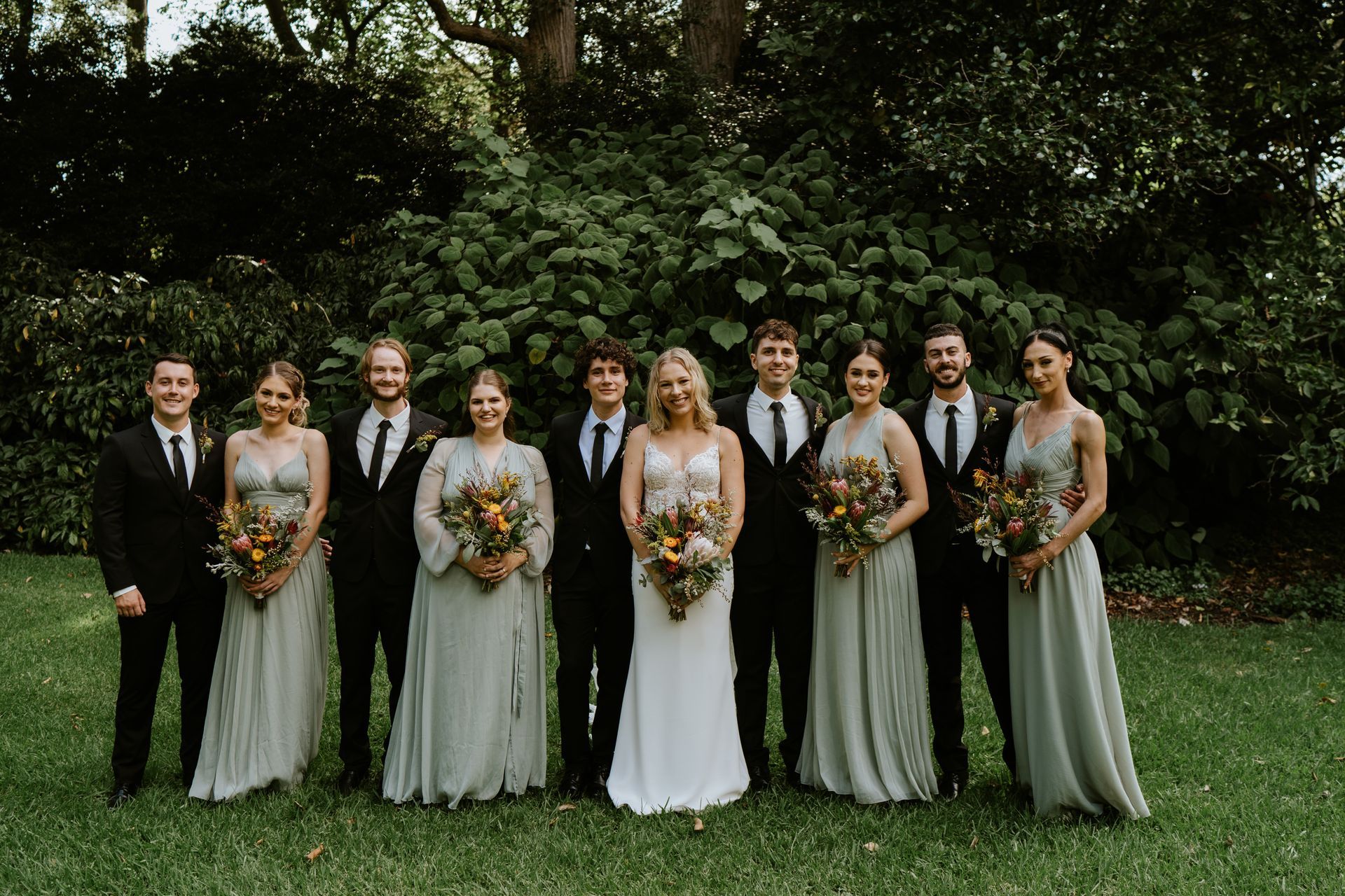Wedding party posing outdoors in front of greenery, holding bouquets and wearing formal attire.