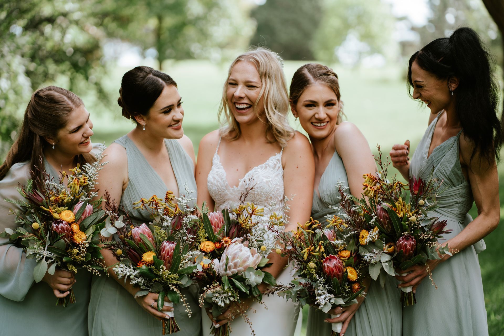 Five women in sage dresses holding colorful wildflower bouquets outdoors, smiling together.