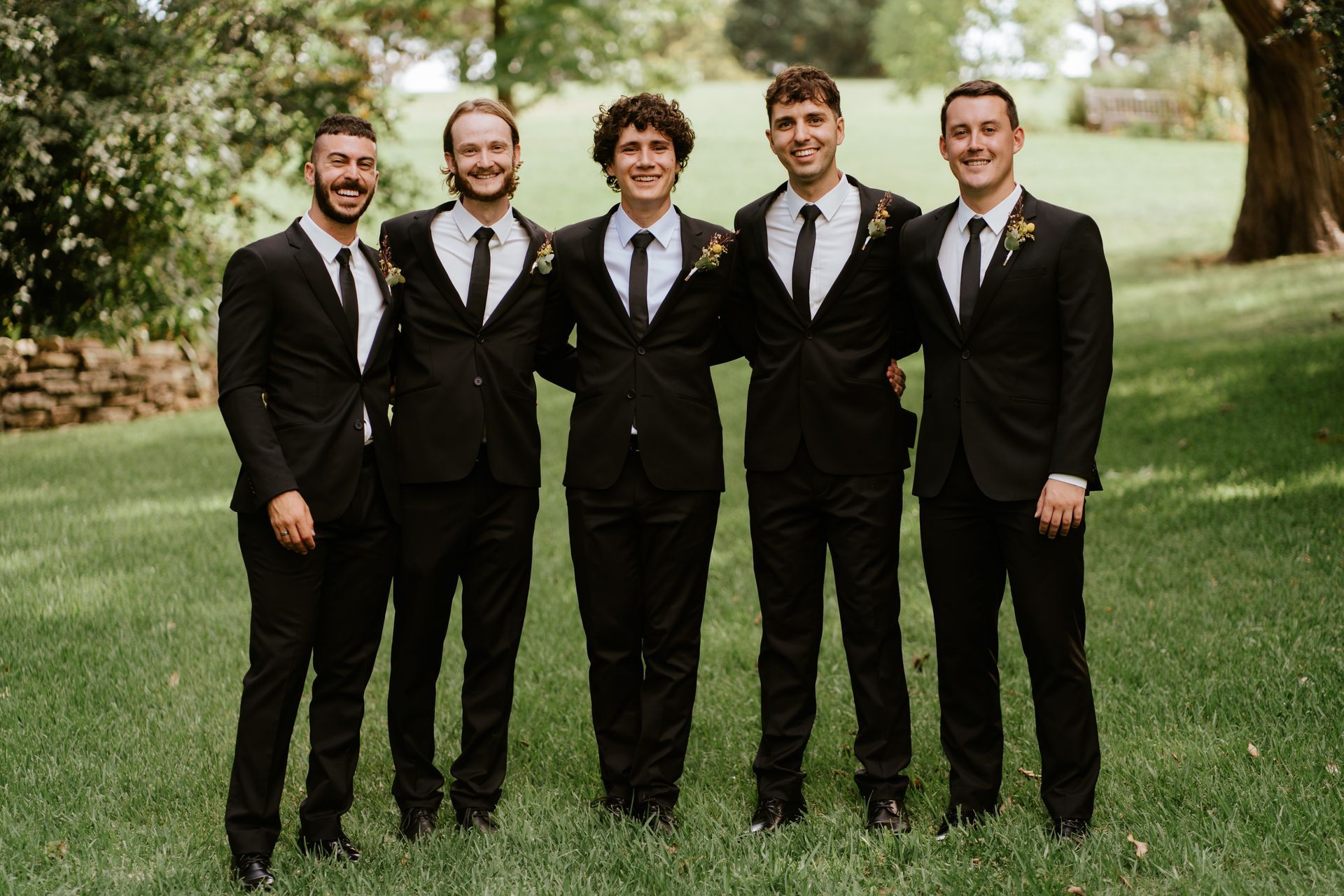 Five men in black suits standing together on grass outdoors, smiling at the camera.