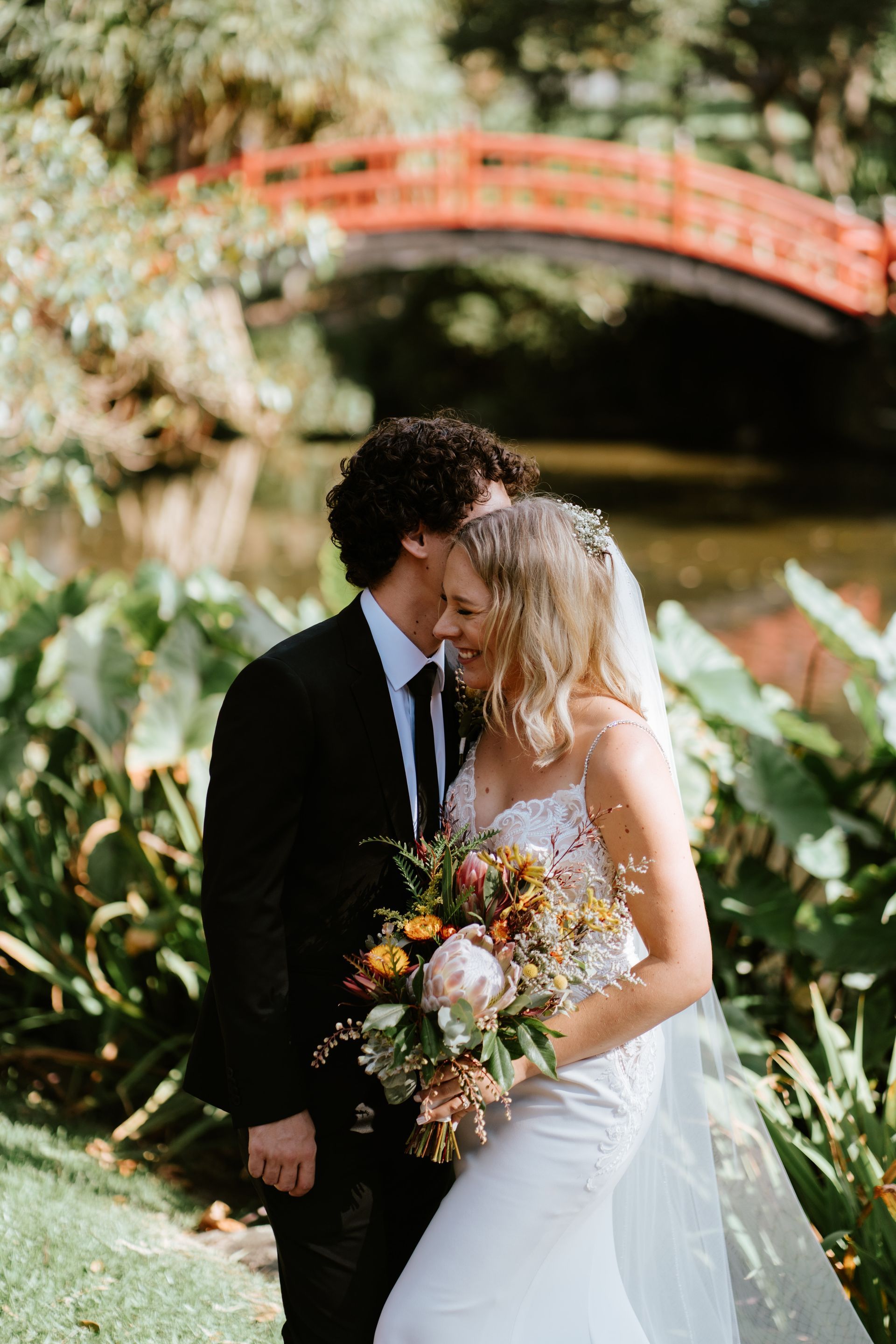 Couple in wedding attire kissing in a garden, holding a bouquet near a red bridge.