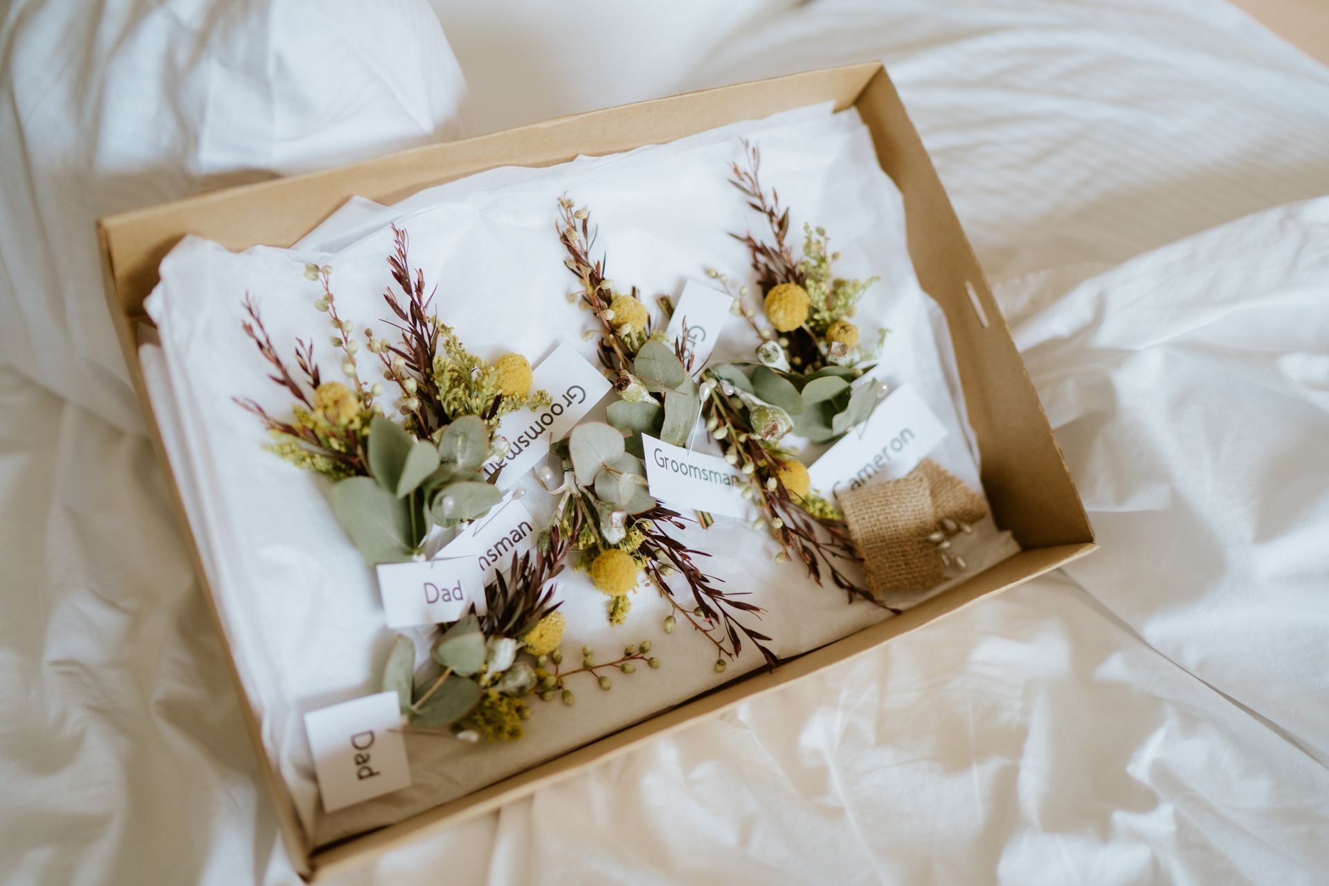 White flower arrangement with dried stems and greenery in a cardboard gift box on a bed
