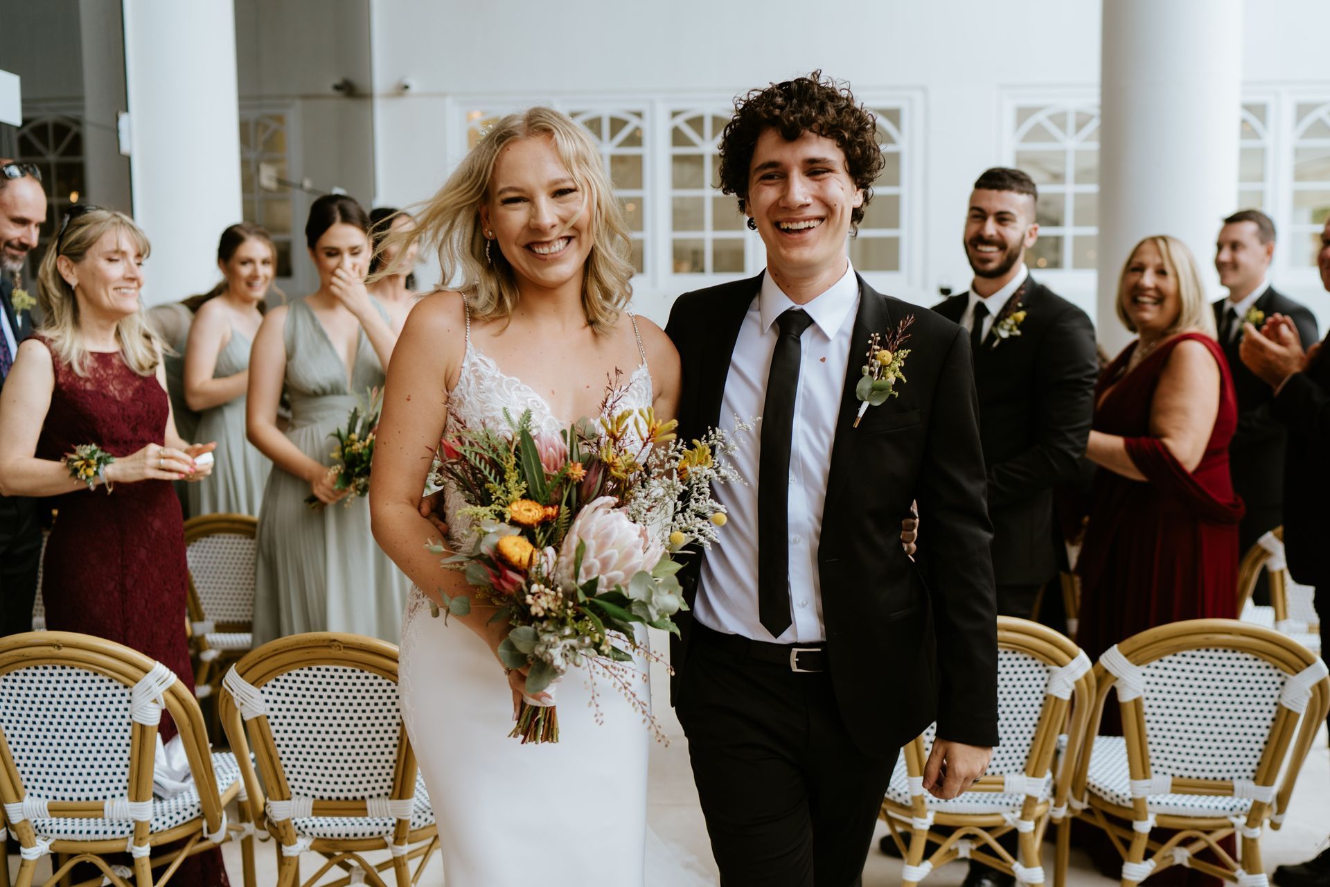 Bride and groom walking down the aisle, smiling, with wedding guests standing behind them.