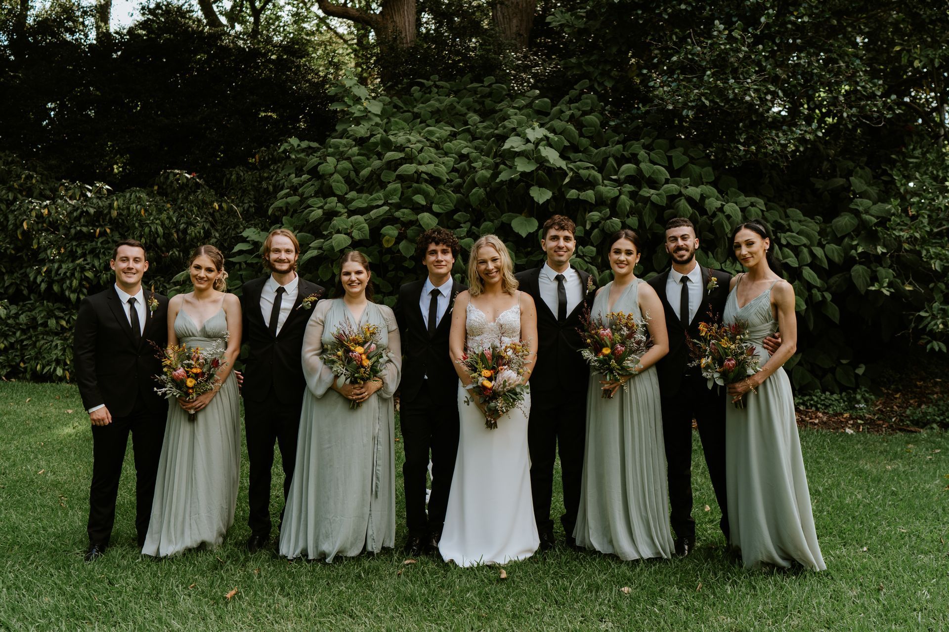 Wedding party posing outdoors on a lawn, holding bouquets, with one person in a black suit and others in light dresses.