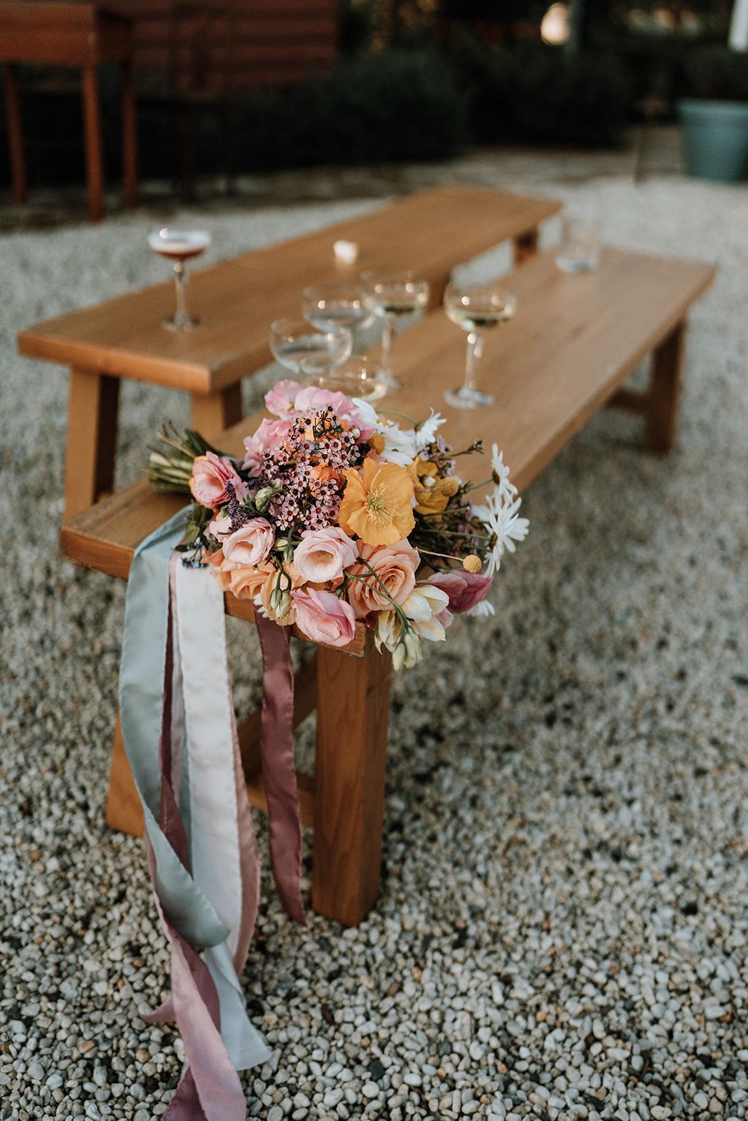 Bouquet with long ribbons on a wooden bench with champagne glasses and a cocktail, outdoors.