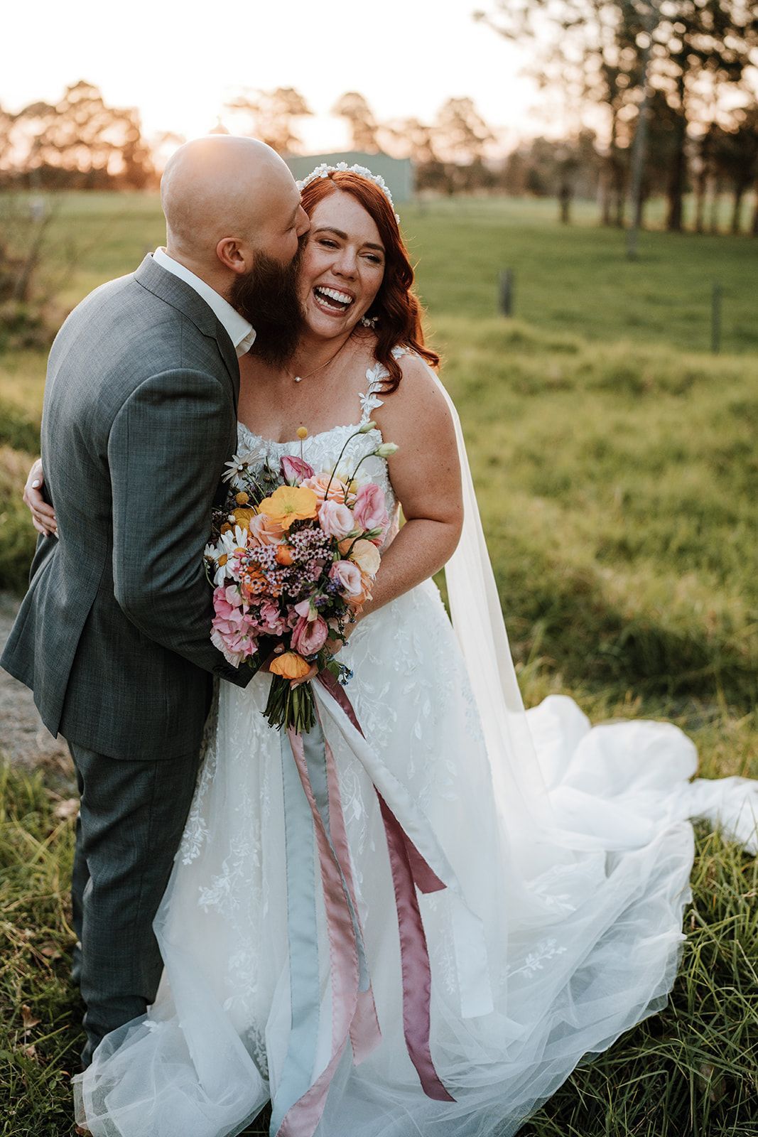 Wedding couple laughing outdoors; groom kissing bride's cheek. Bride in white dress, bouquet; sunny field background.