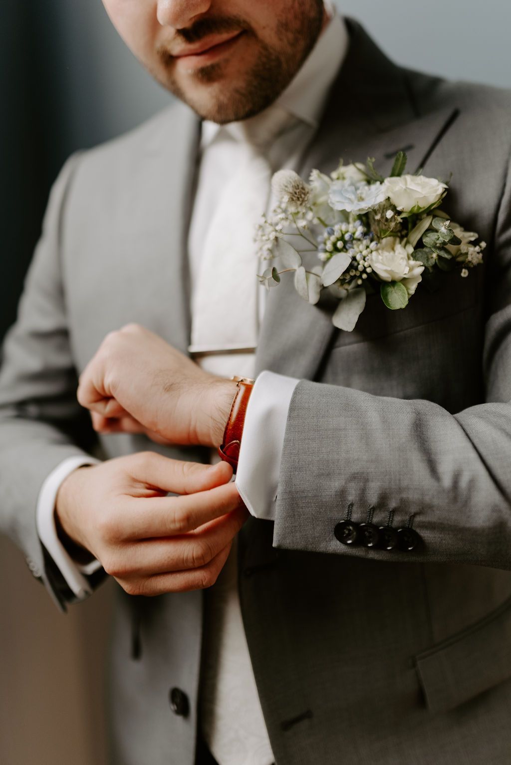 Man in a gray suit fastening a red bracelet beside a white floral boutonniere