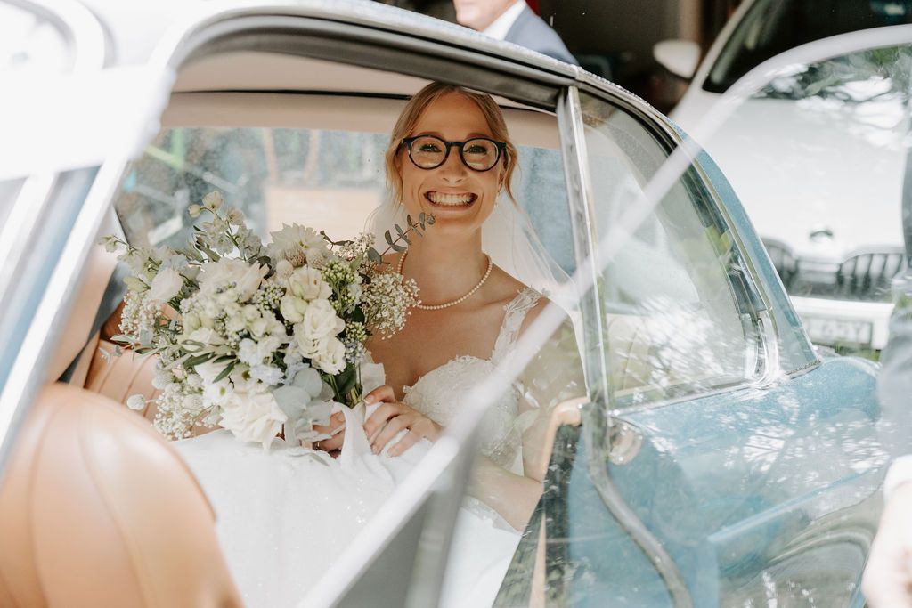 Smiling bride in a white dress holding a bouquet inside a car, viewed through the open door
