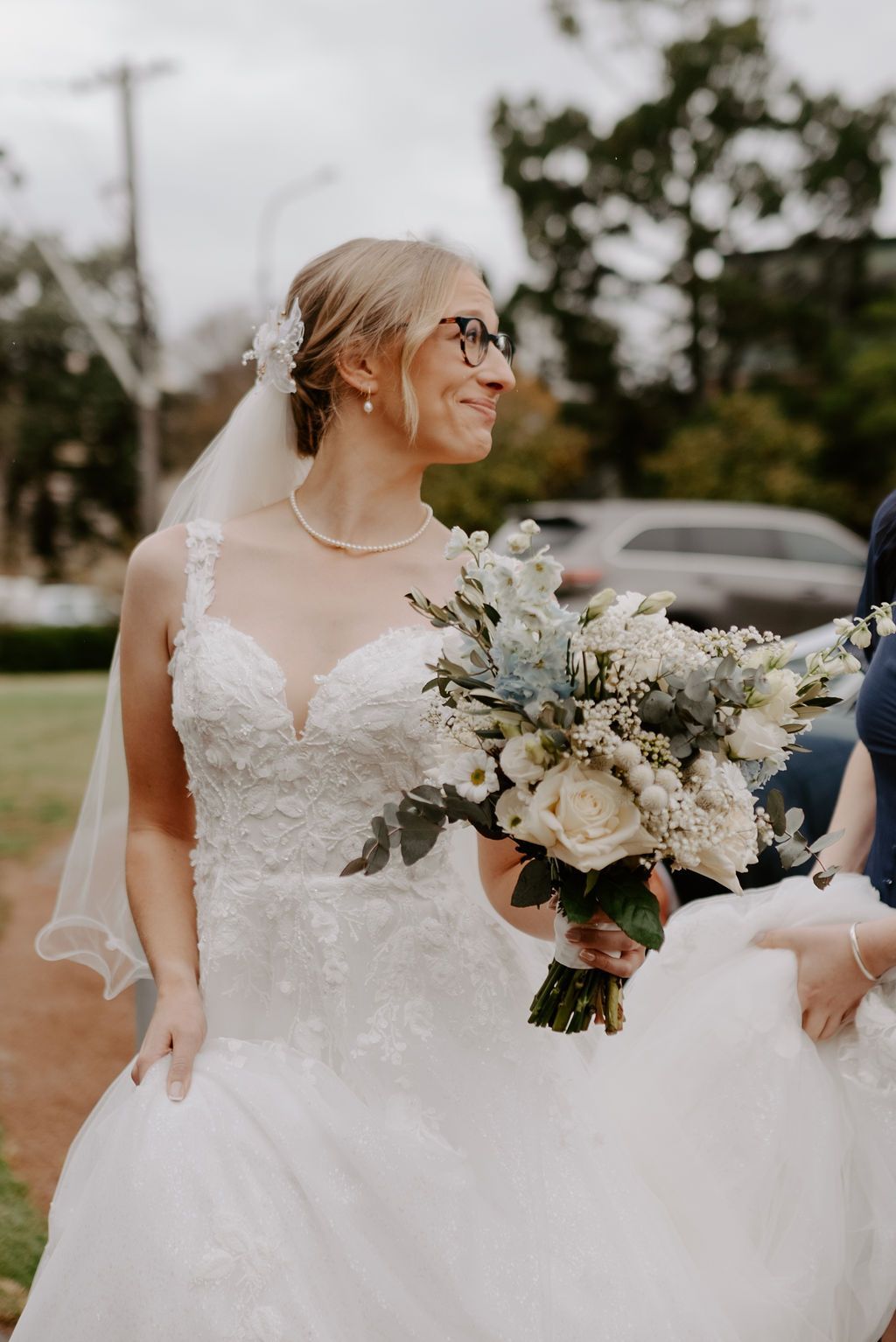 Bride in white lace gown holding bouquet outdoors, smiling beside a car
