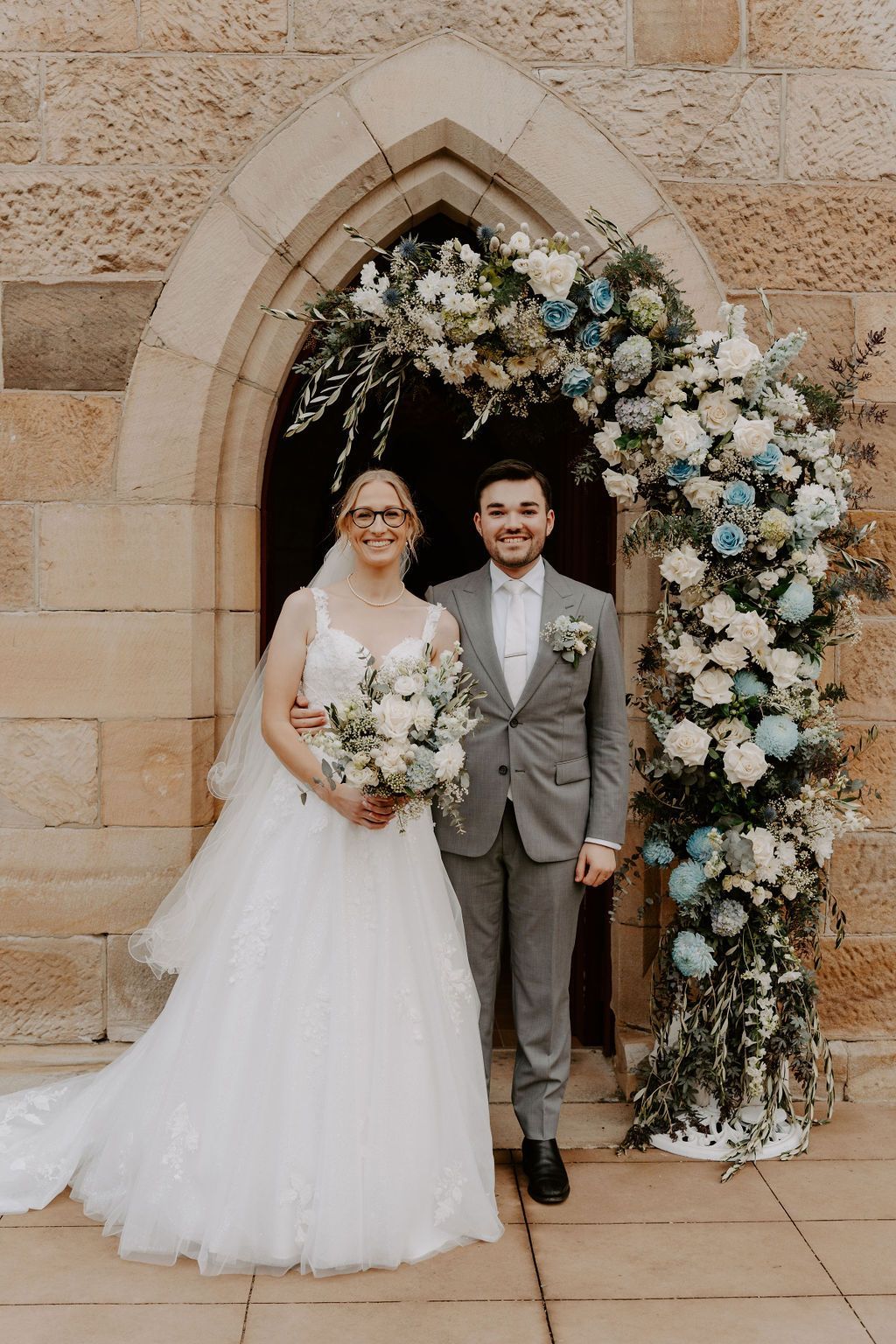 Bride and groom posing under a floral arch beside a stone chapel entrance