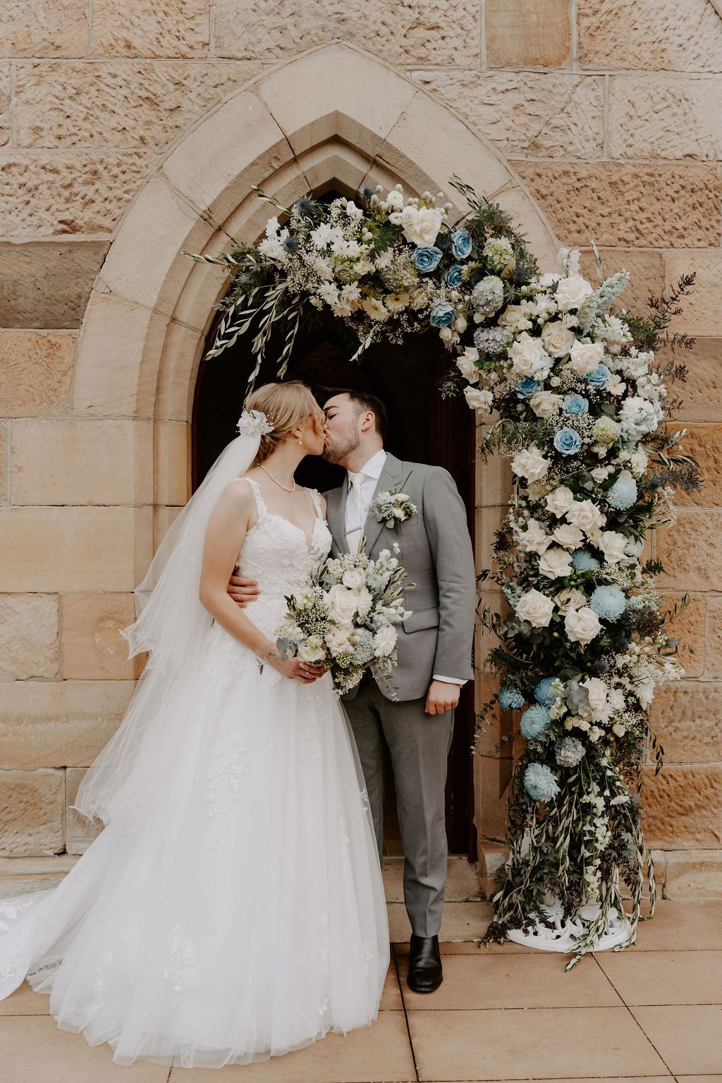 Bride and groom kissing under a floral arch outside a stone building