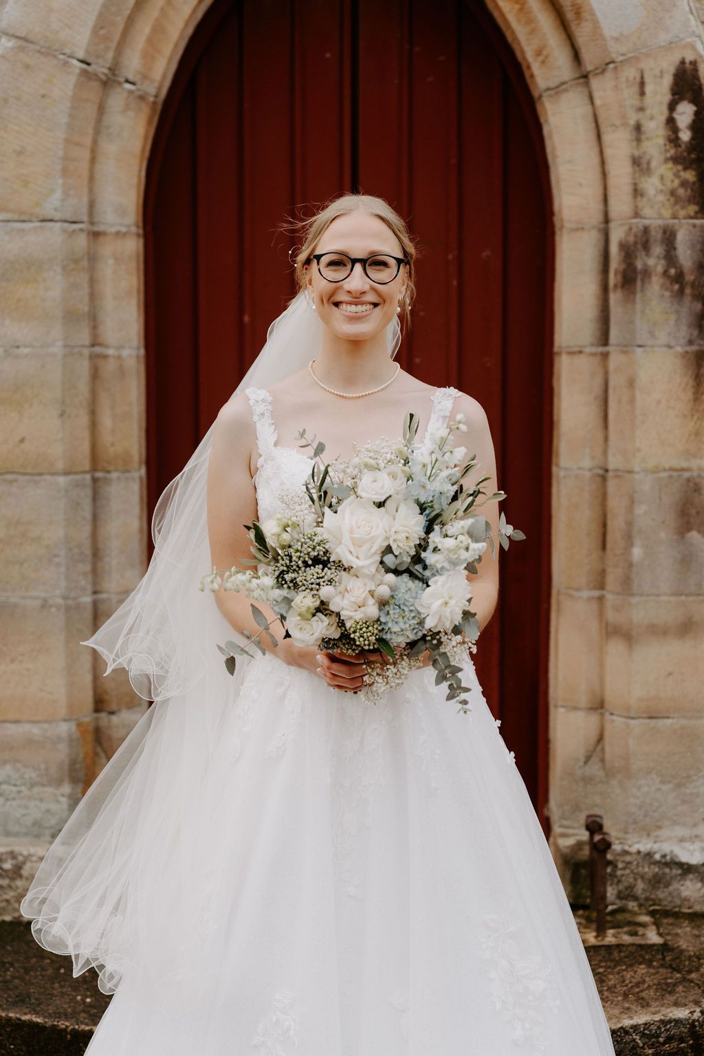 Bride in a white gown holding a bouquet, standing before a red church doorway.
