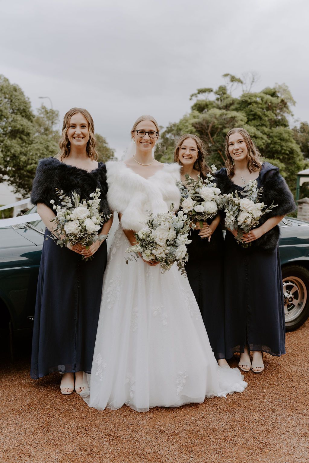 Four women in formal dresses holding bouquets, standing outdoors in front of a car.