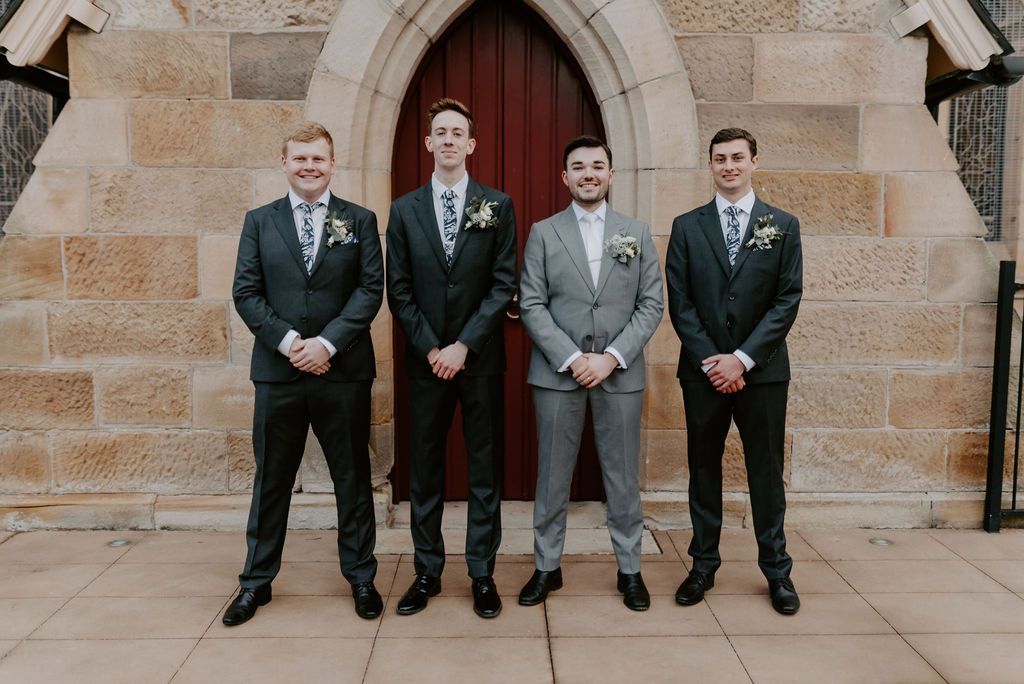 Four men in suits standing in front of a stone archway, hands clasped, posing for a formal photo.