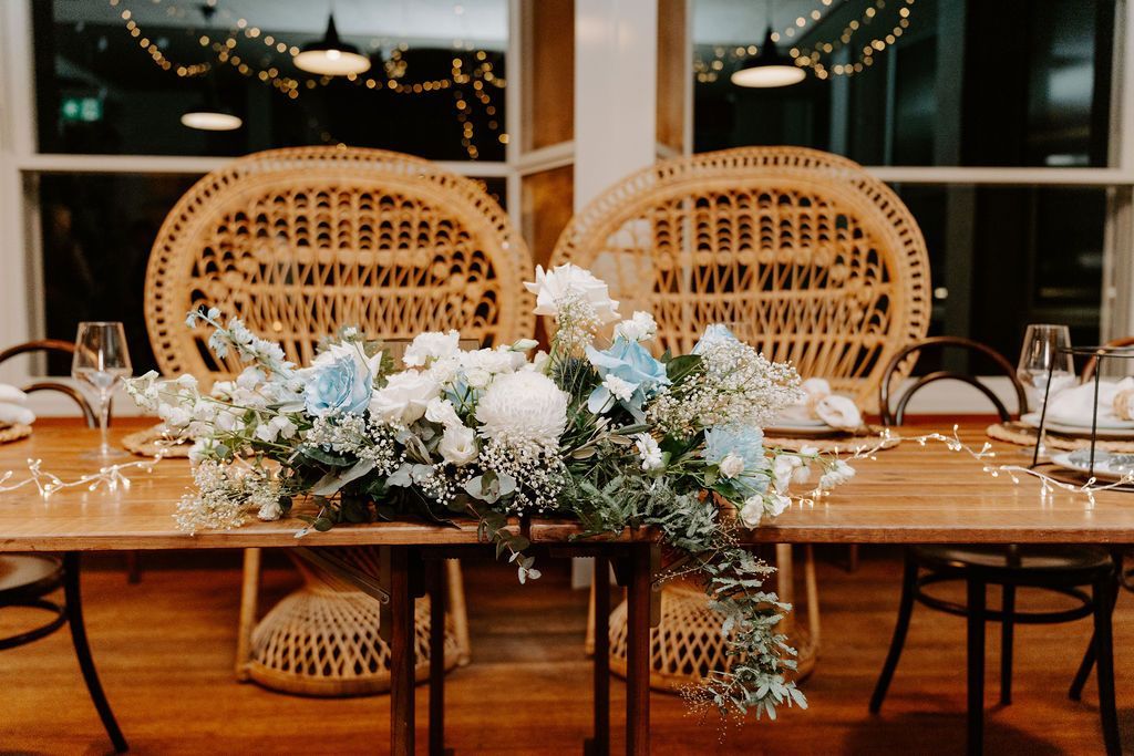 Floral wedding reception table with two wicker chairs and string lights in a cozy indoor venue