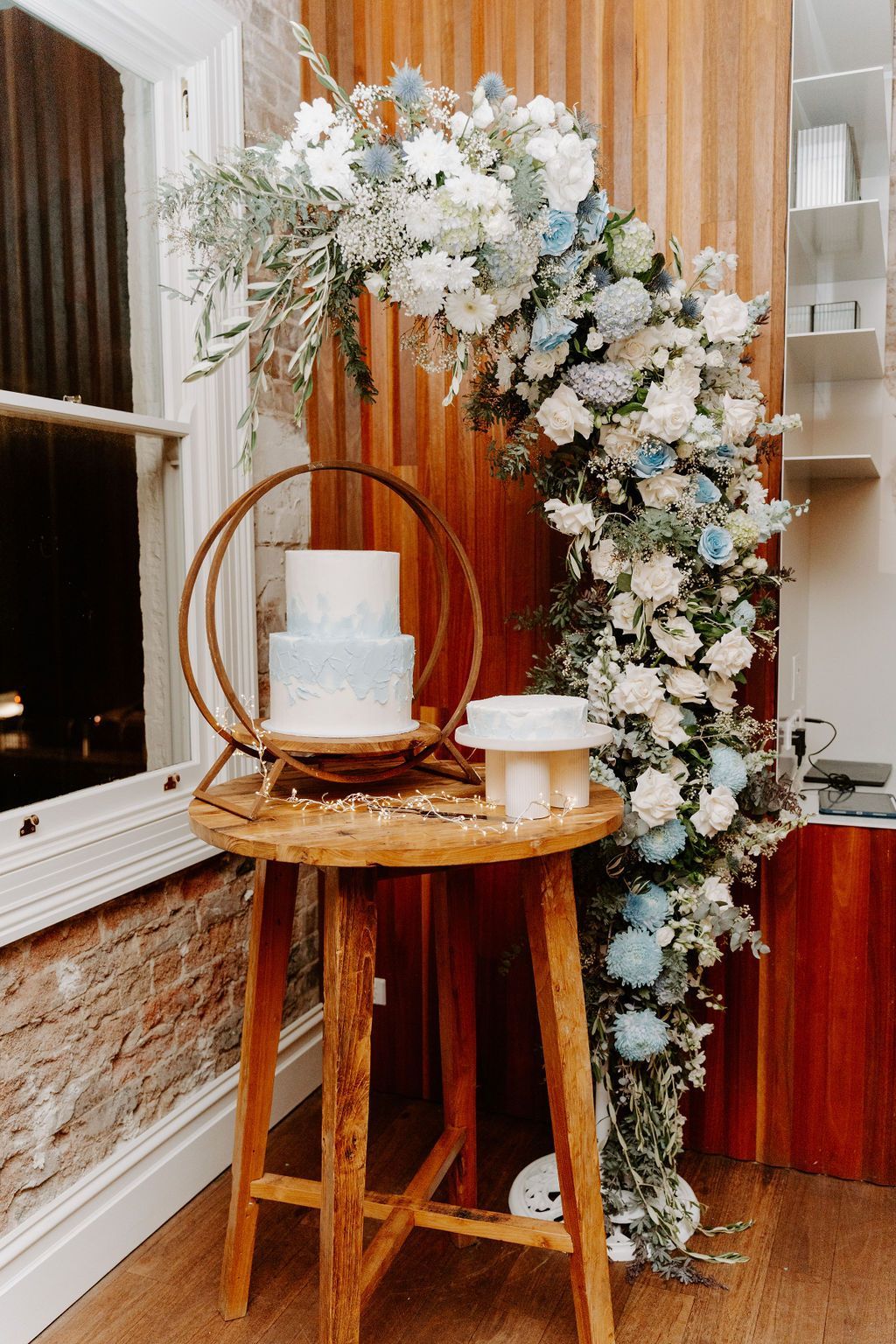 Elegant floral arch with white and blue flowers beside a small table holding a cake and candles