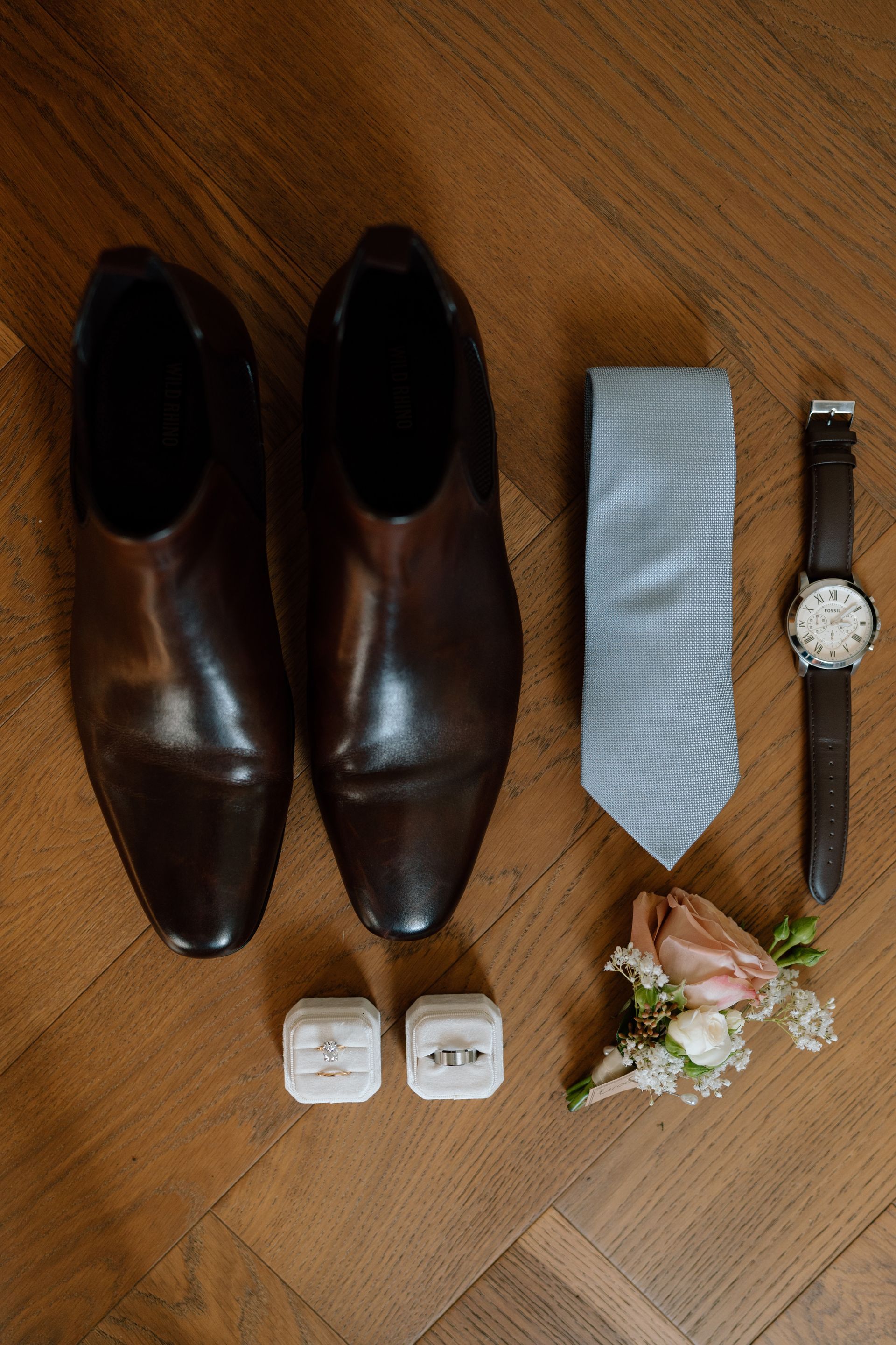Brown dress shoes, light blue tie, wristwatch, cufflinks, and boutonniere arranged on a wooden floor.
