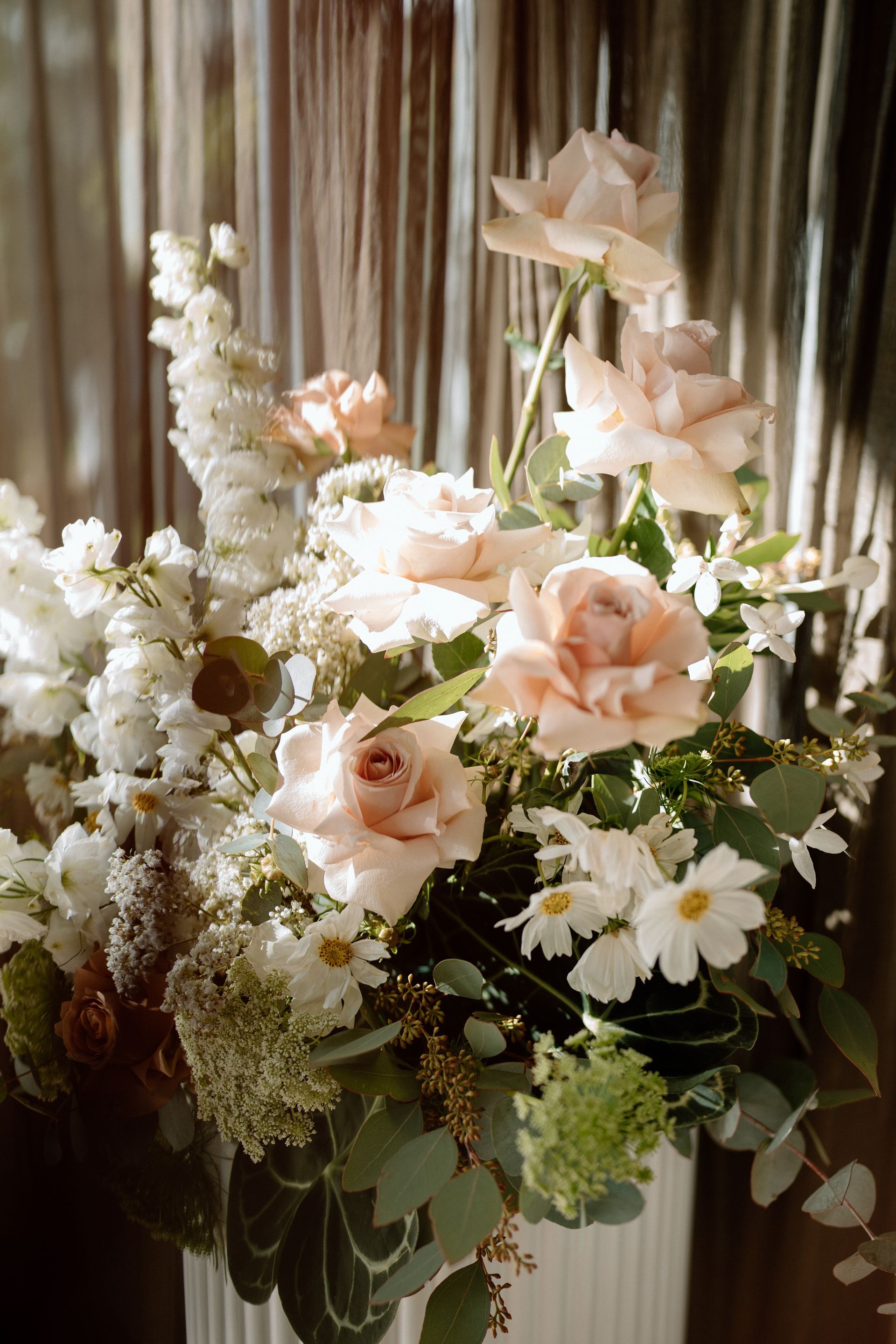 Elegant pastel floral arrangement with white daisies, pale pink roses, and greenery against draped curtains