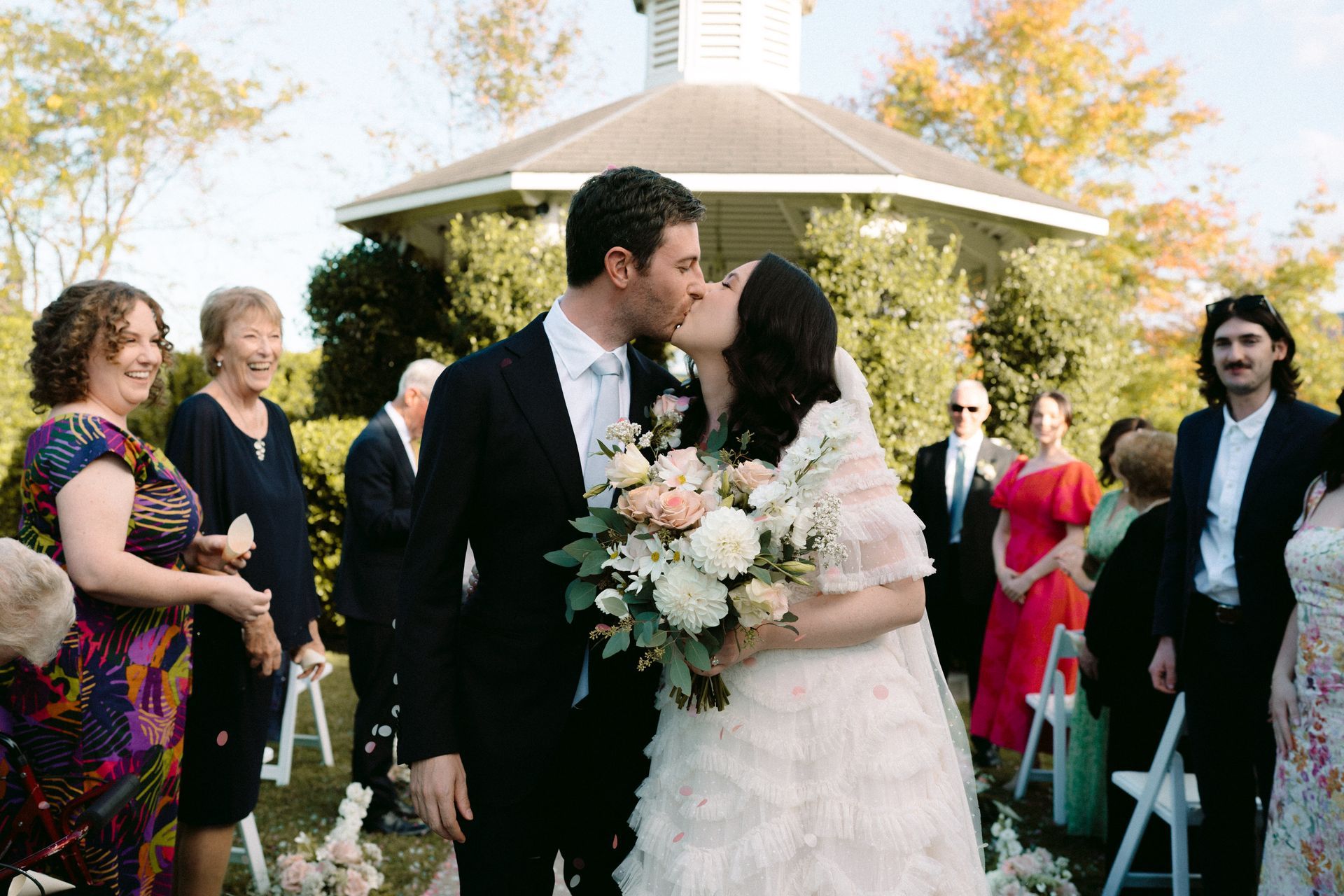 Bride and groom kiss at an outdoor wedding, with guests standing nearby under a gazebo.
